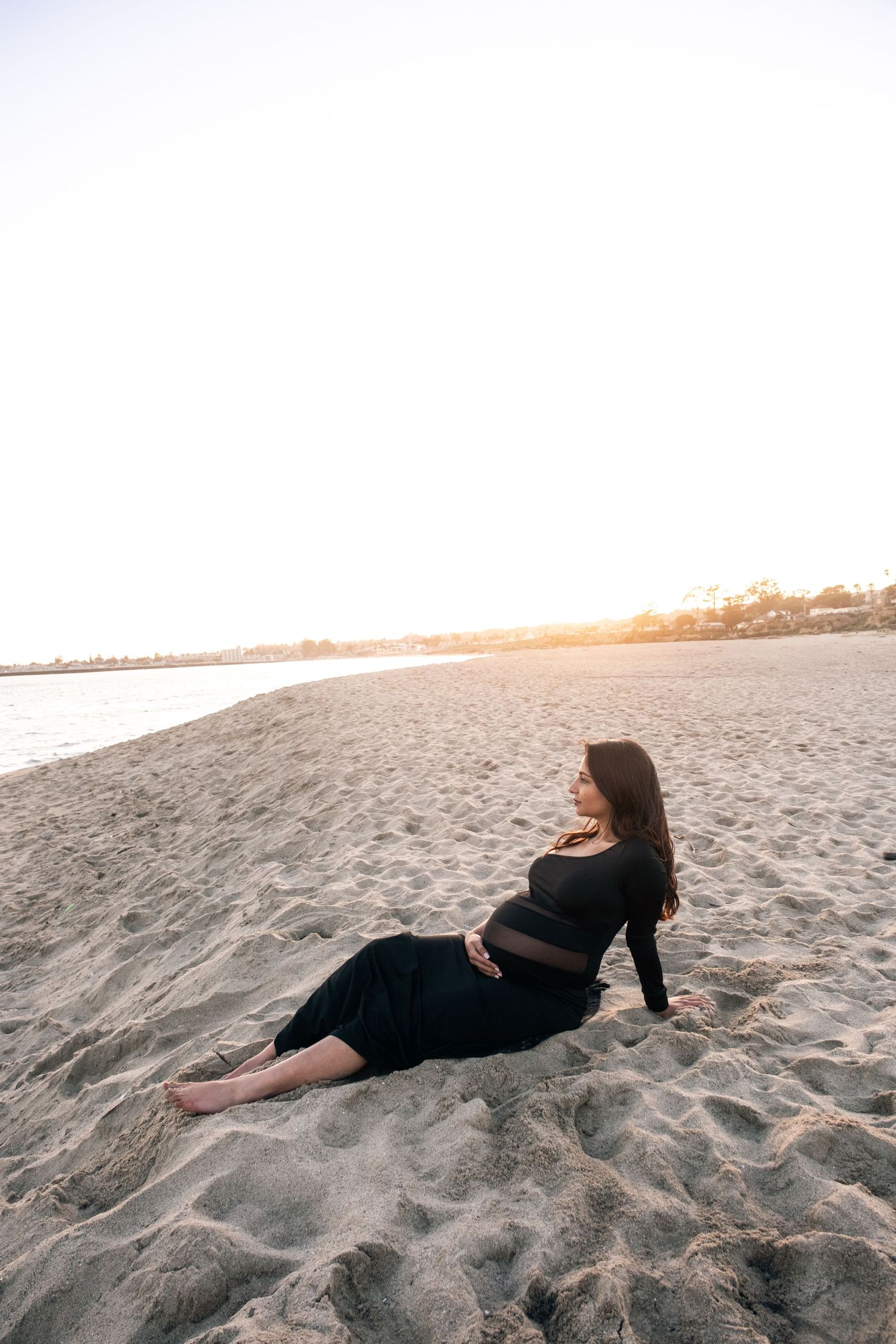 Artistic maternity beach photo with subject lying in sand at golden hour.