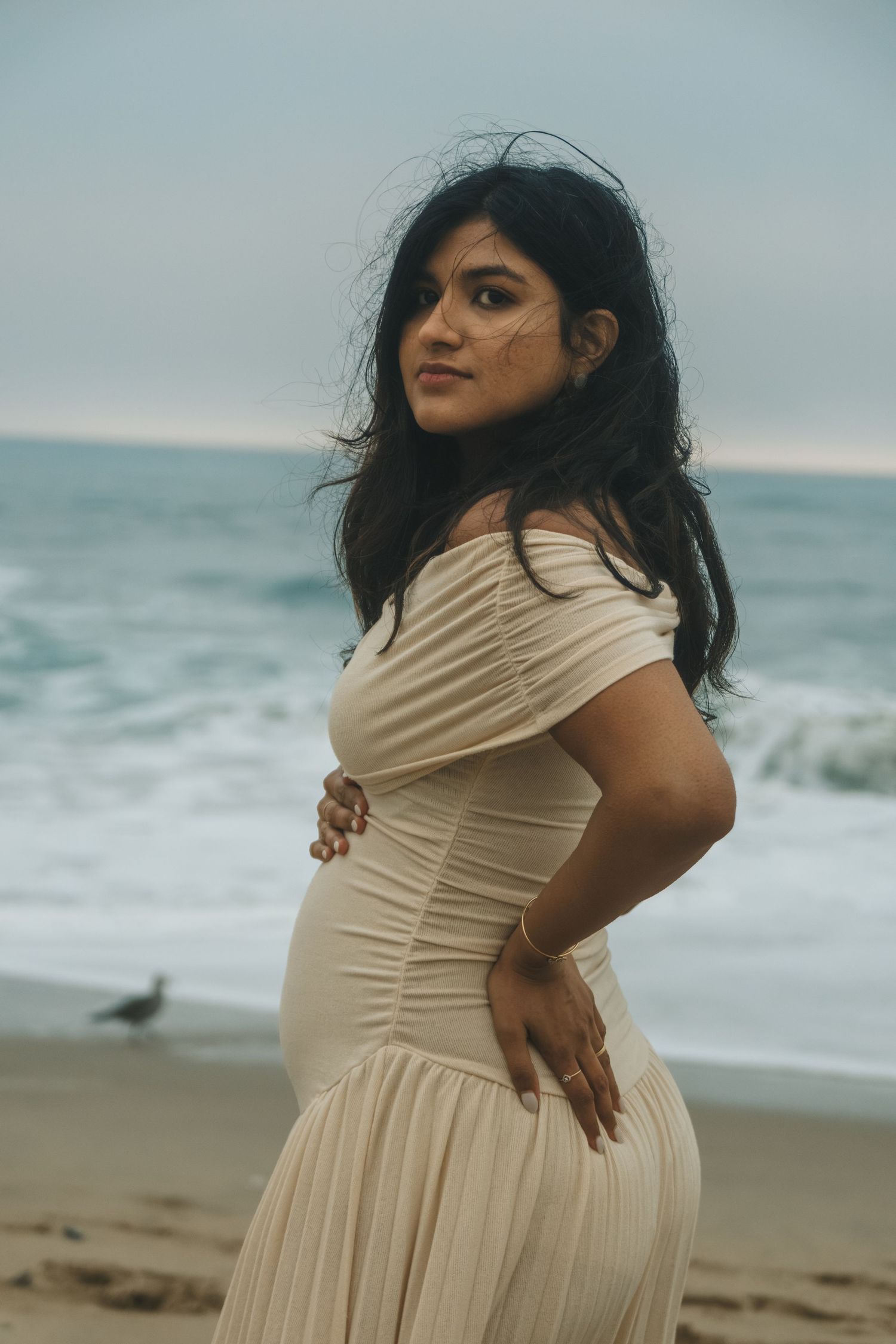 A person in a flowing cream dress poses on a beach with waves in the background.