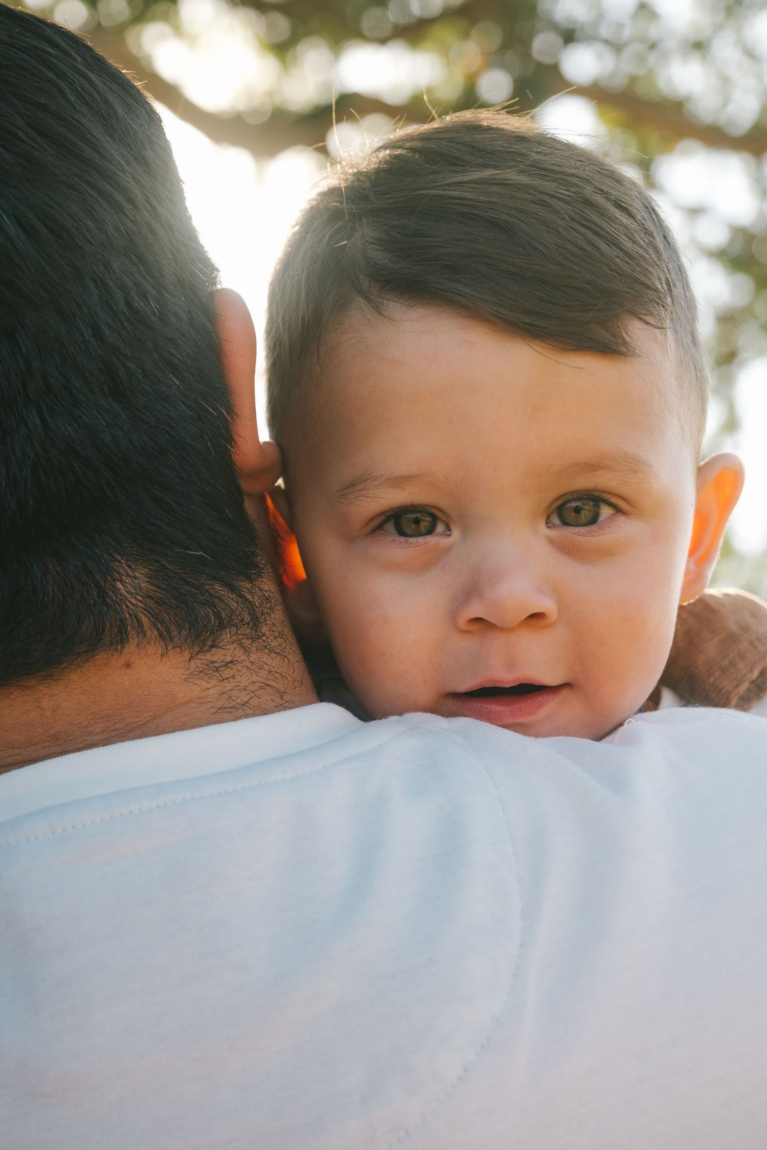 Close-up shot of a baby's smiling face peeking over a shoulder in sunlight.