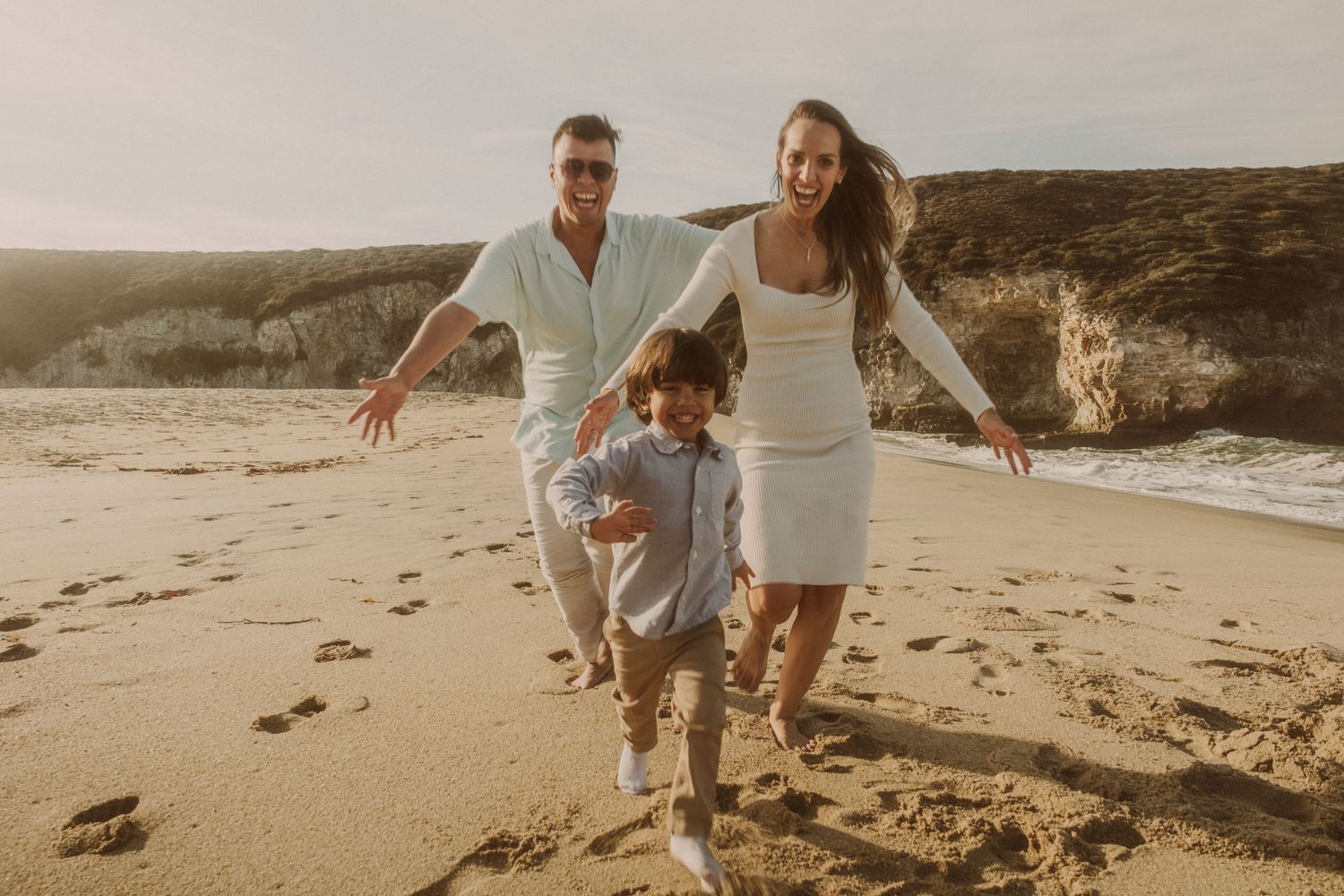 Family running together on a sandy beach during sunset with cliffs in background.