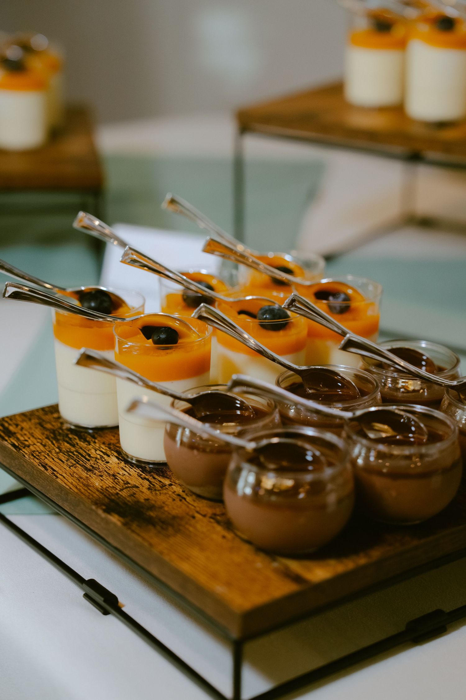 Elegant dessert display with chocolate mousse, peach panna cotta, and vanilla cups with spoons on a wooden tray.