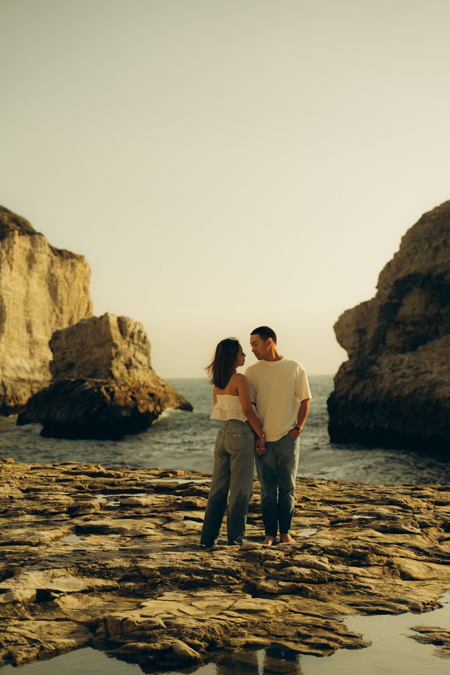 Couple holding hands on rocky coastal shore at golden hour, framed by dramatic cliffs with ocean view.