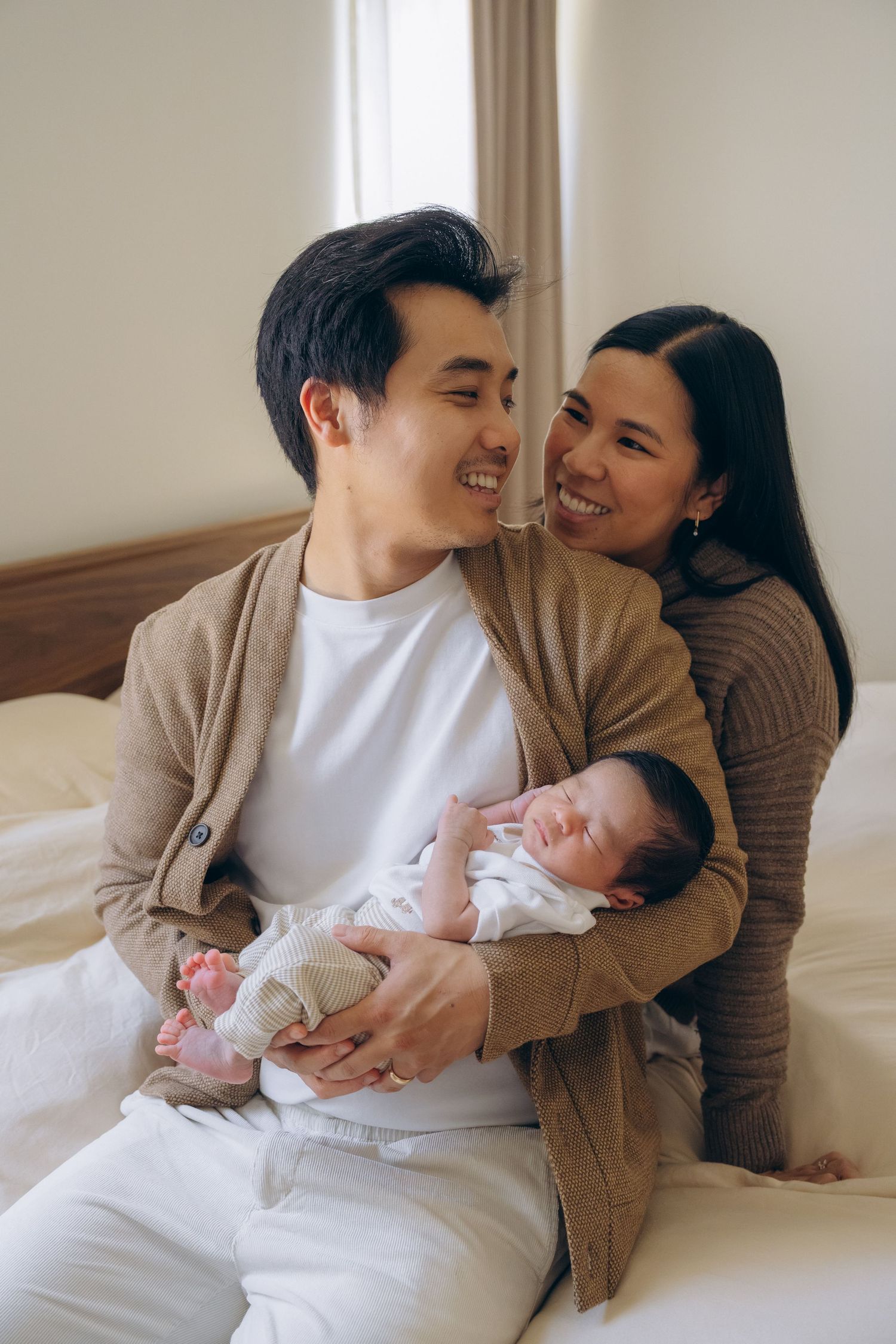 Happy Asian couple sitting on bed smiling at each other while father holds sleeping newborn baby.