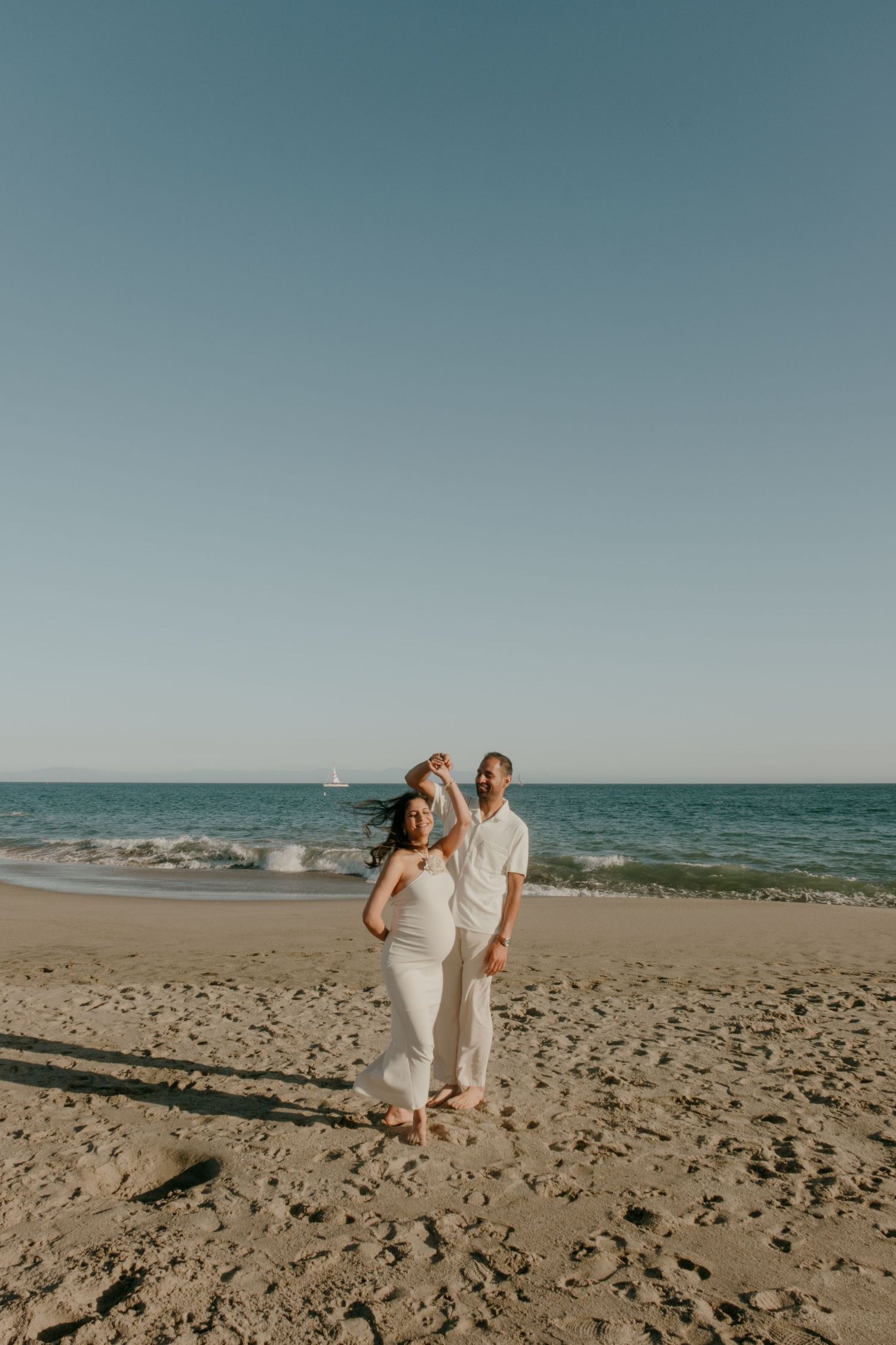 Couple walking hand in hand on a sunny beach with waves in the background.