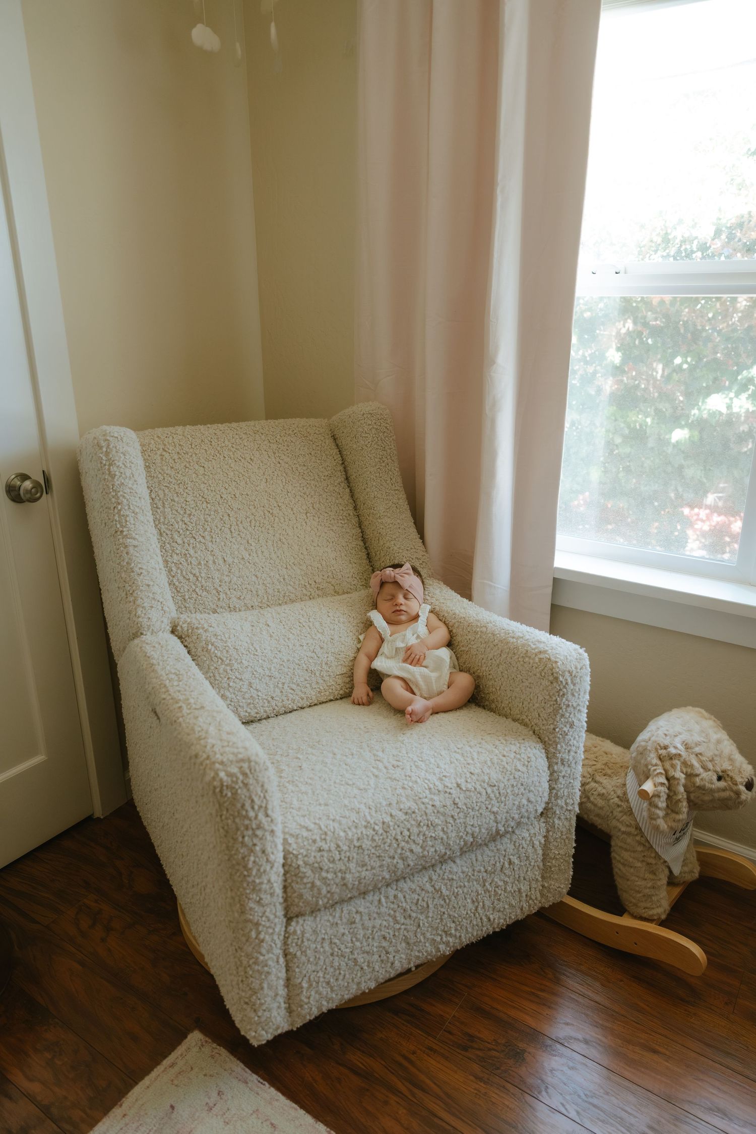 A newborn baby in a floral outfit sits in a cream boucle rocking chair beside a bright window in a nursery.