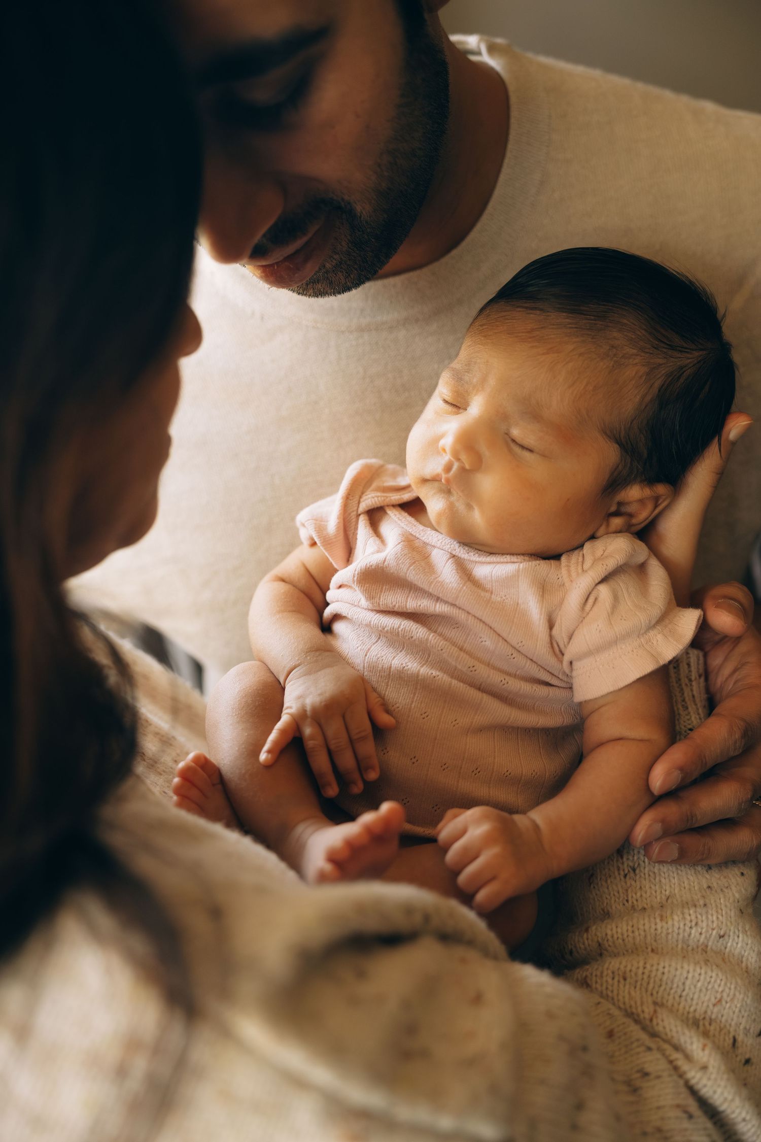 Newborn baby in pink onesie sleeping peacefully in fathers arms while mother gazes lovingly at the infant.