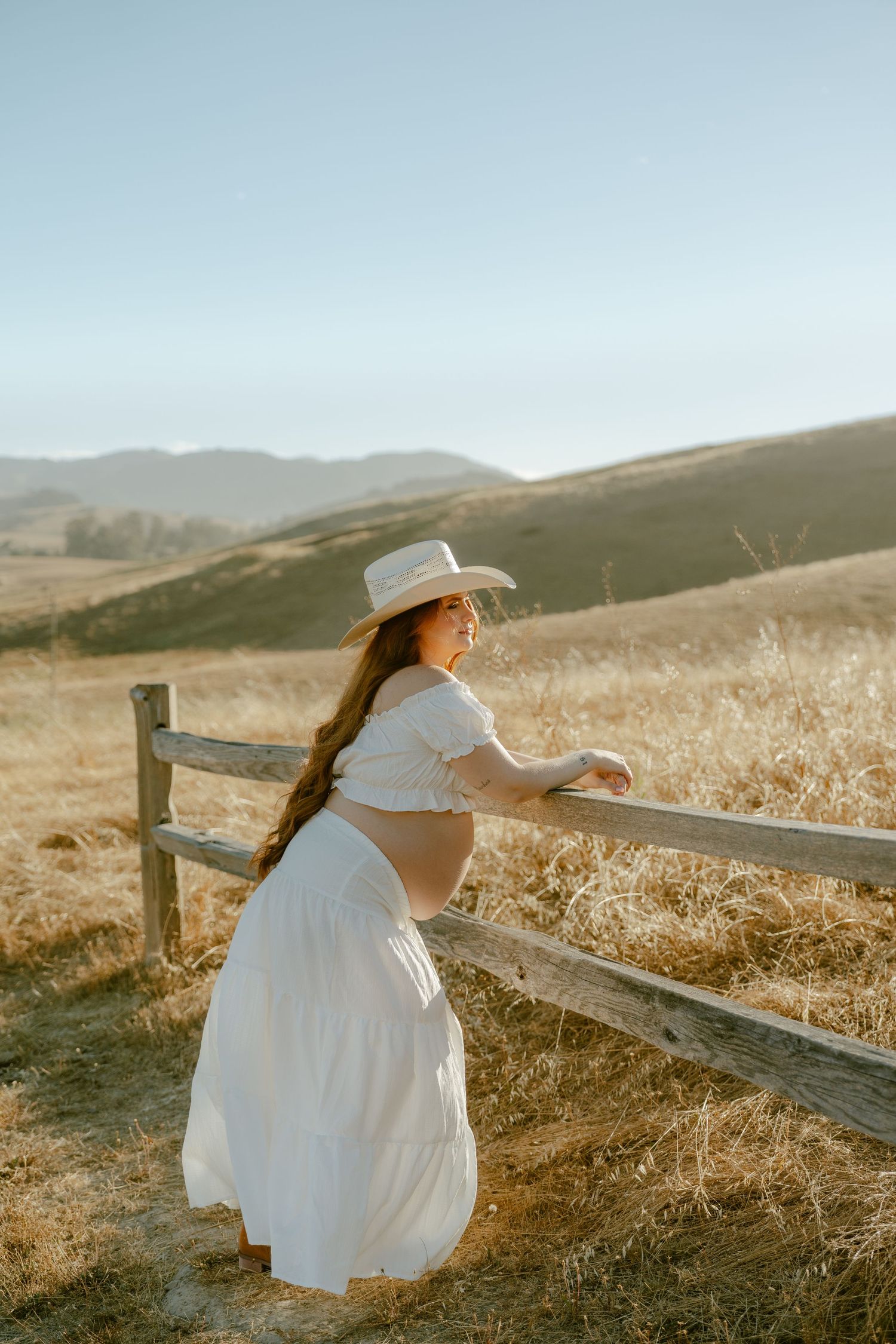 Pregnant woman in white crop top, skirt, and cowboy hat leaning on a wooden fence in a golden field with rolling hills.