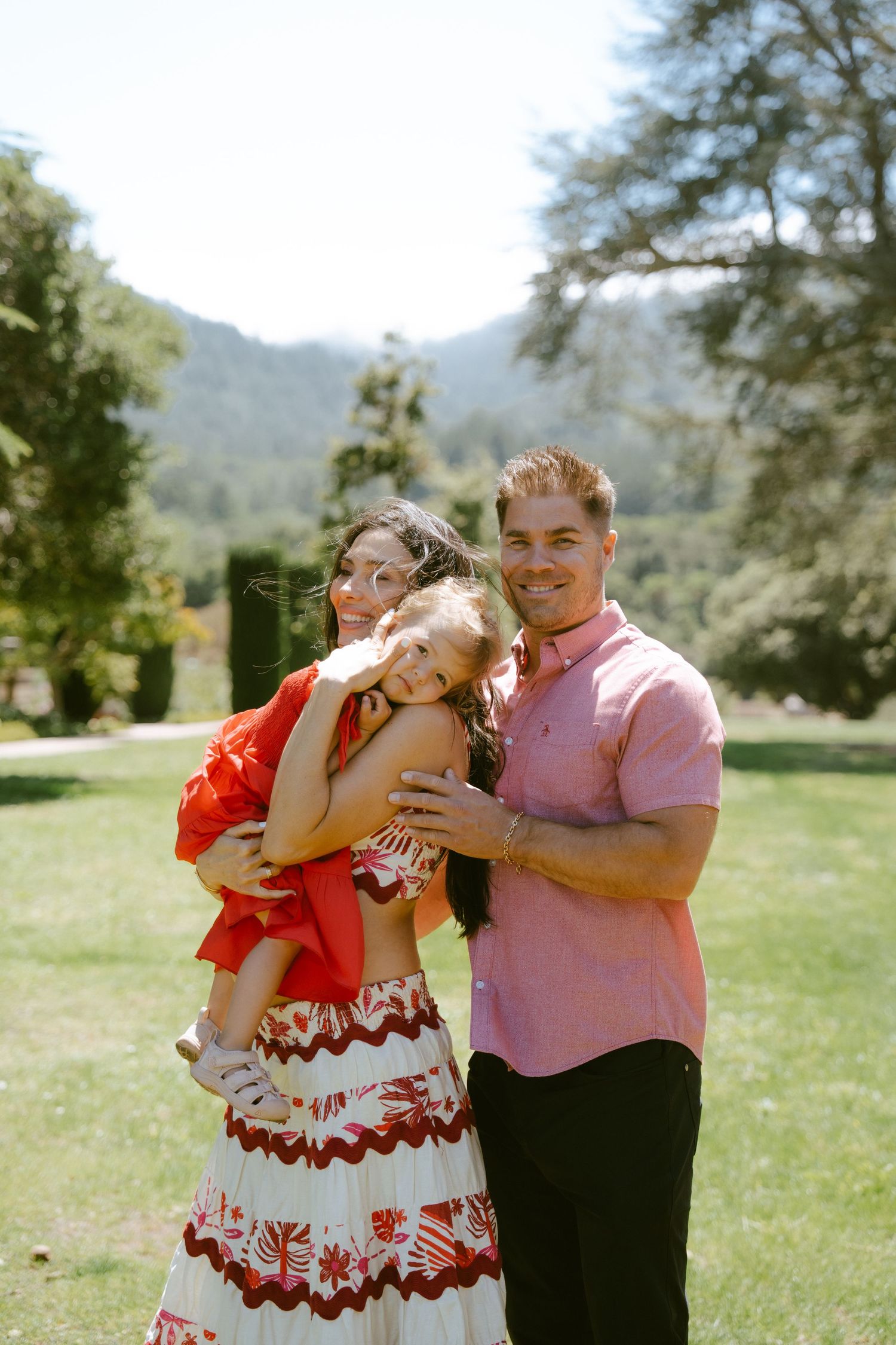 Happy family of three posing outdoors on green lawn with mountain backdrop on a sunny day.