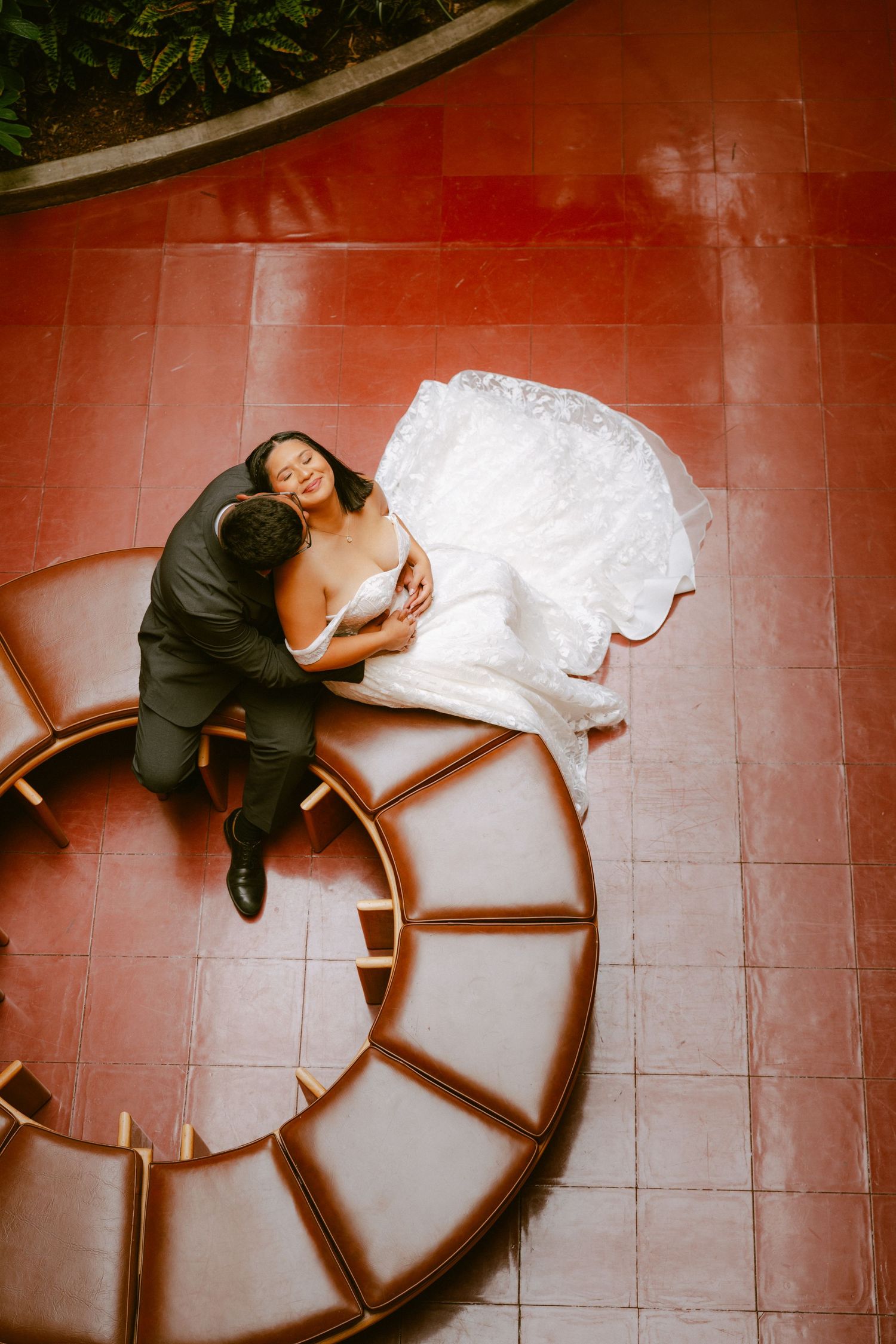 Aerial view of bride and groom posing on circular staircase, bride in lace gown, groom in black suit.