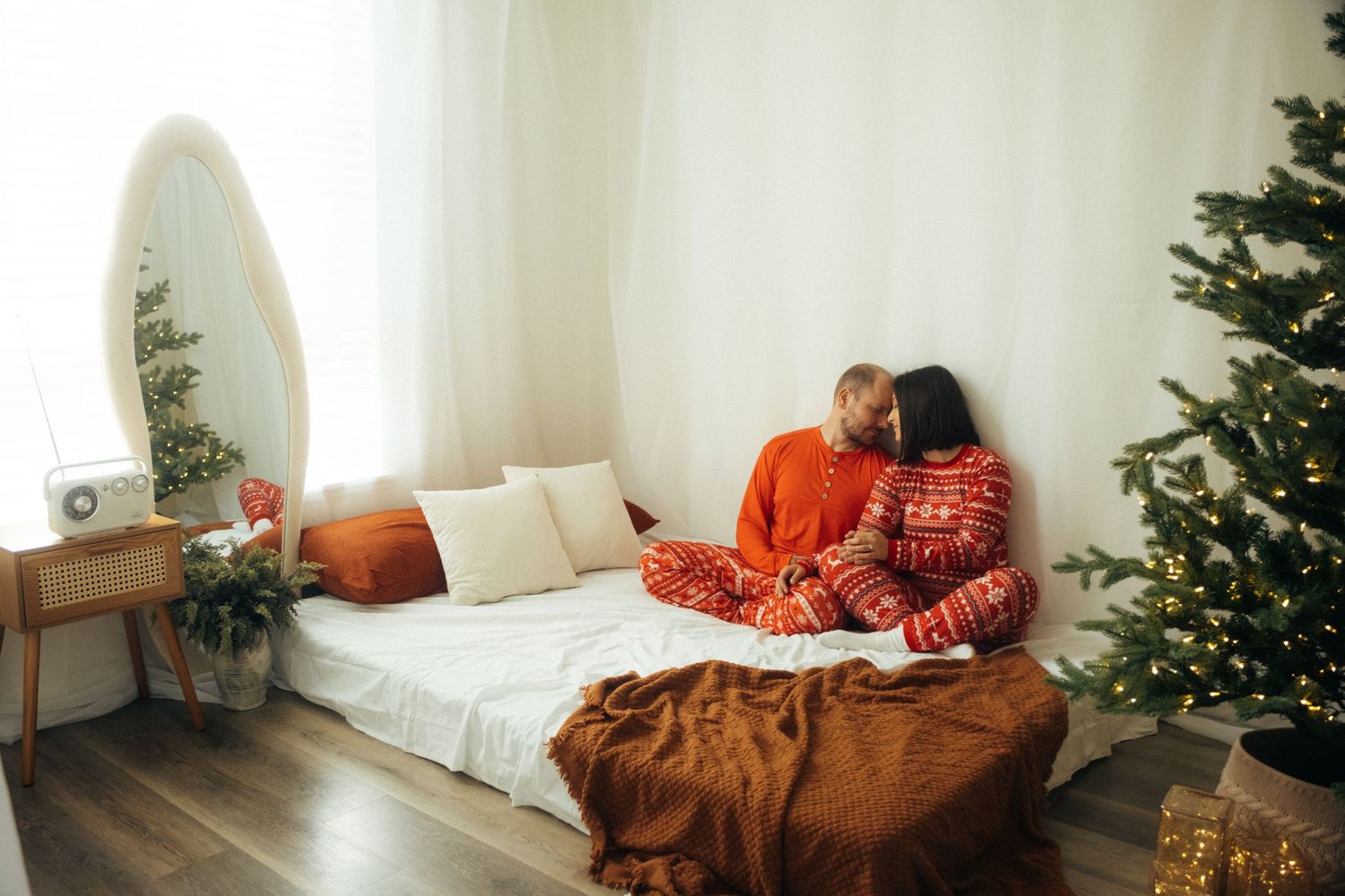 Couple in coordinating red holiday outfits cuddling on a white mattress near a Christmas tree.