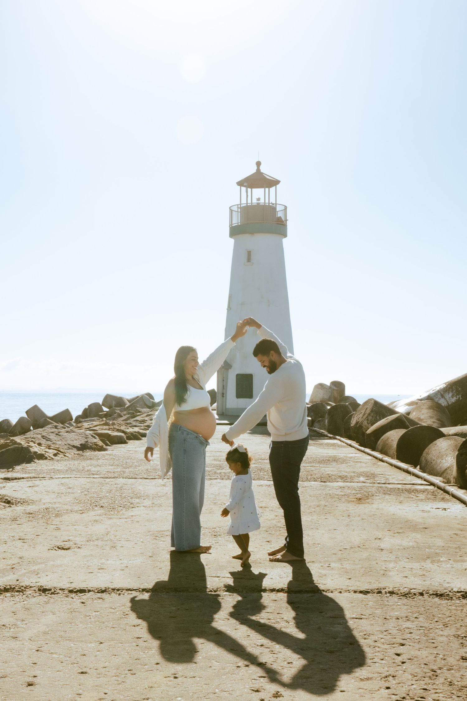 Family silhouettes stand near a lighthouse on a rocky beach during a bright sunny day.