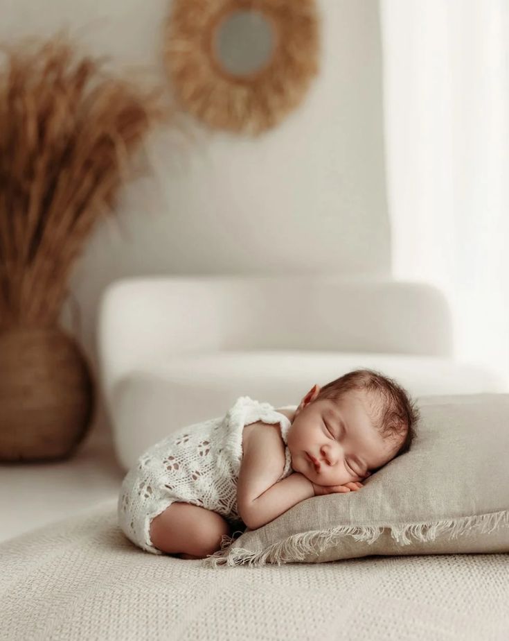 A sleeping newborn baby wearing white lace lies peacefully on a beige cushion in a minimalist setting.