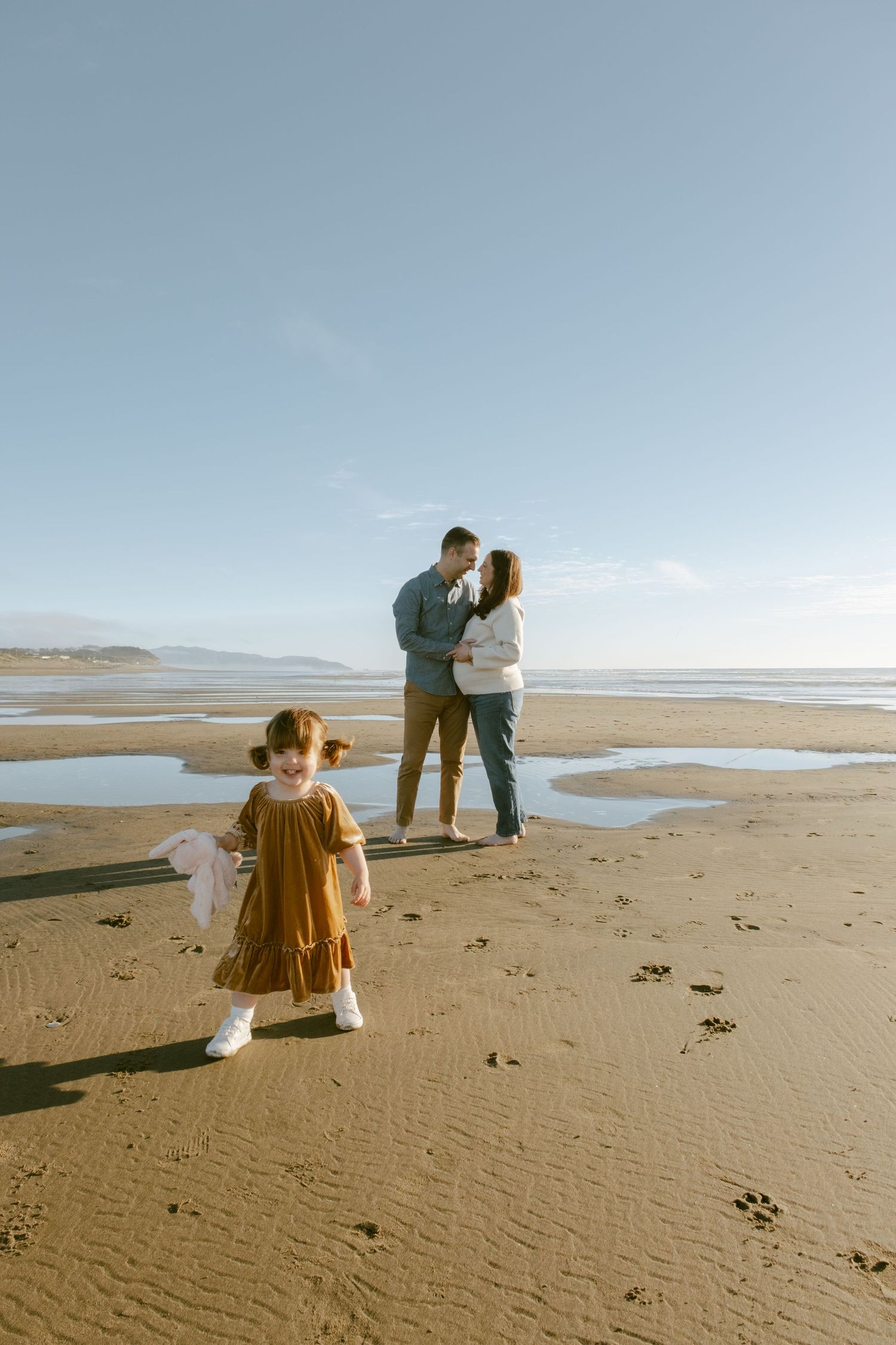 A young girl in a brown dress walks on a sandy beach while her parents share a romantic moment by the ocean.