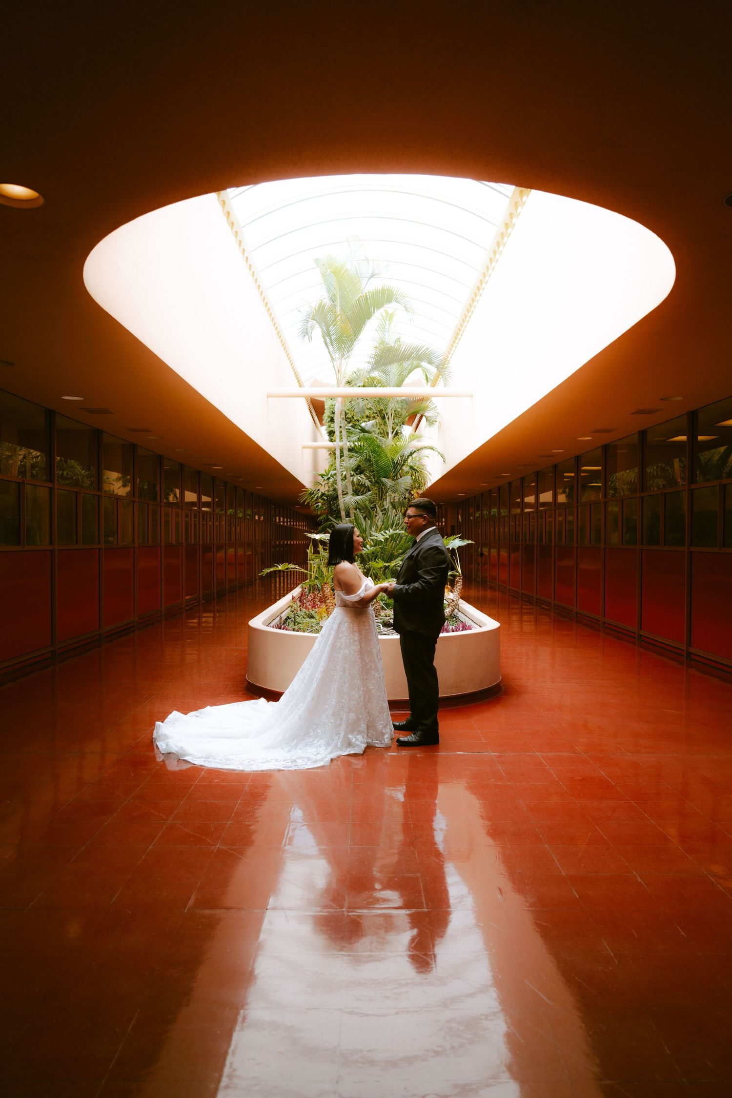 Bride and groom share a romantic moment in a stunning red corridor with a triangular skylight and tropical plants.