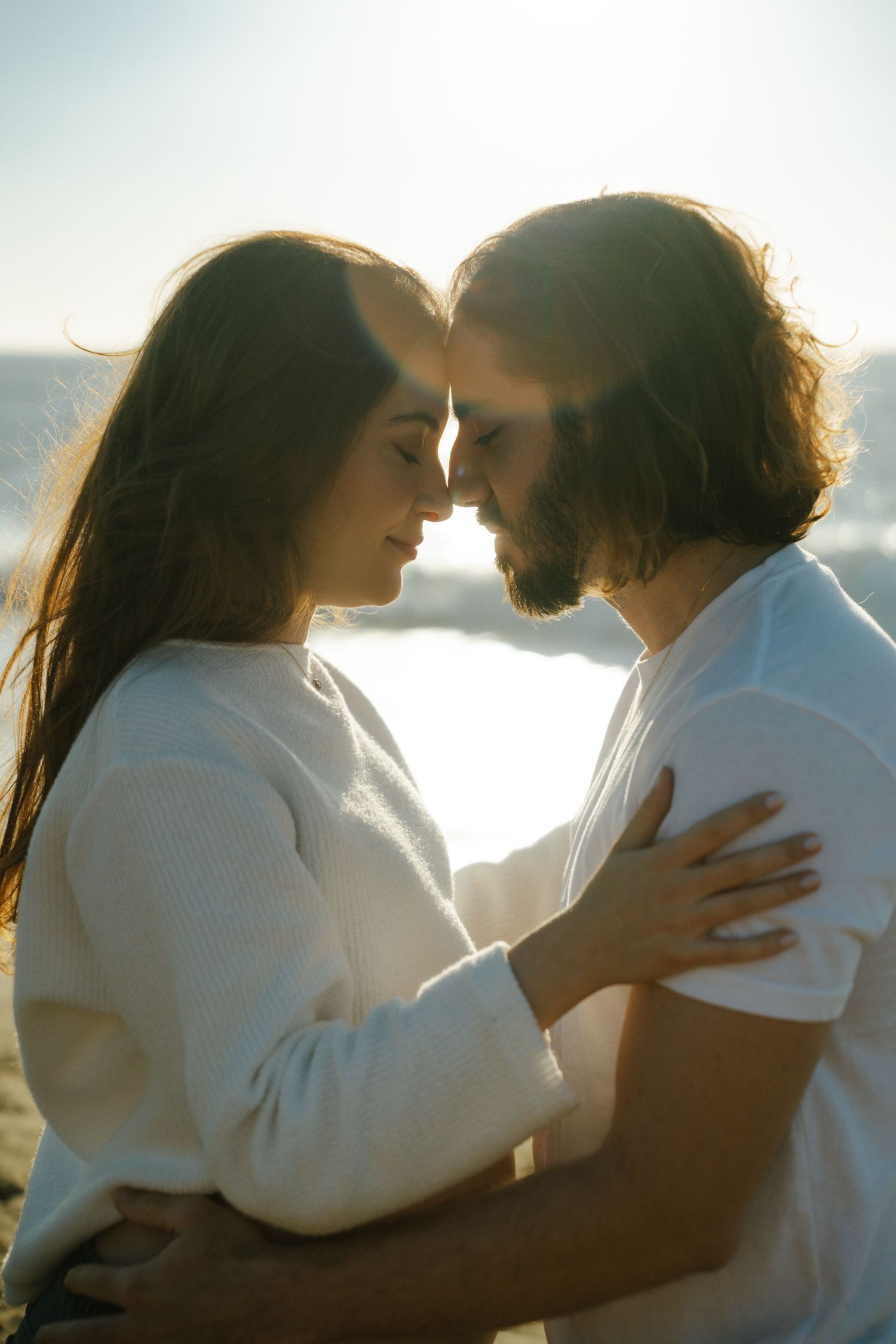 Romantic couple in white embracing at sunset beach, foreheads touching, backlit by golden sunlight near ocean.