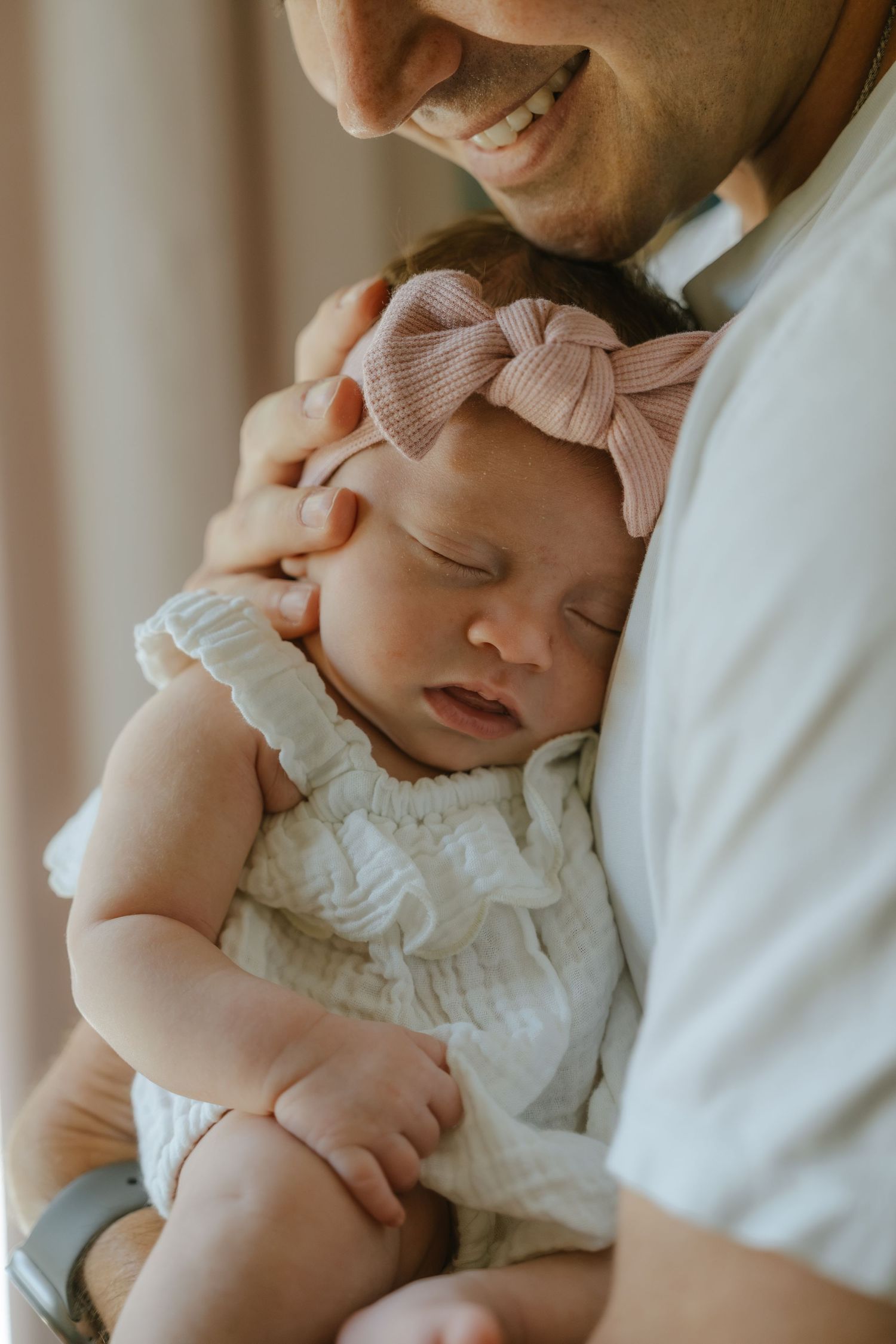 Smiling father cradling sleeping newborn baby girl wearing white outfit and pink bow headband.