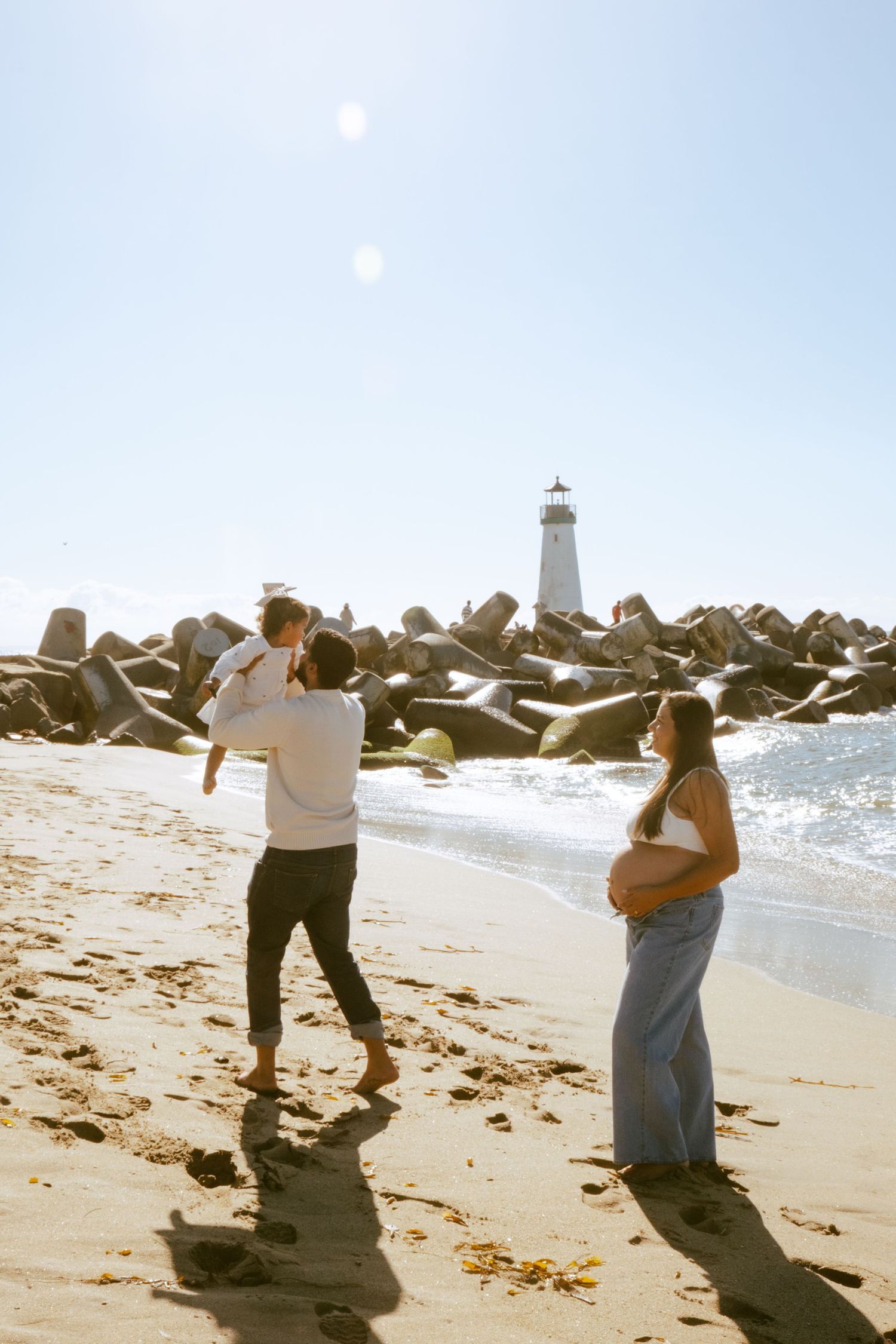 Family on sunny beach near lighthouse, pregnant mother watches father lift child near rocky breakwater.