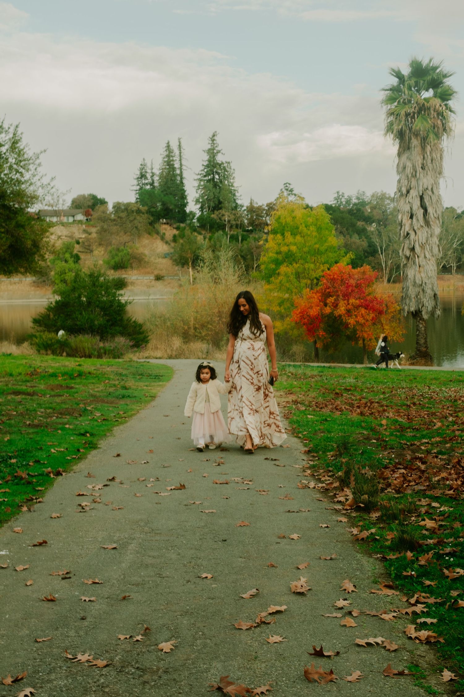 A scenic autumn path with fallen leaves where two people in flowing dresses walk together in a park setting.