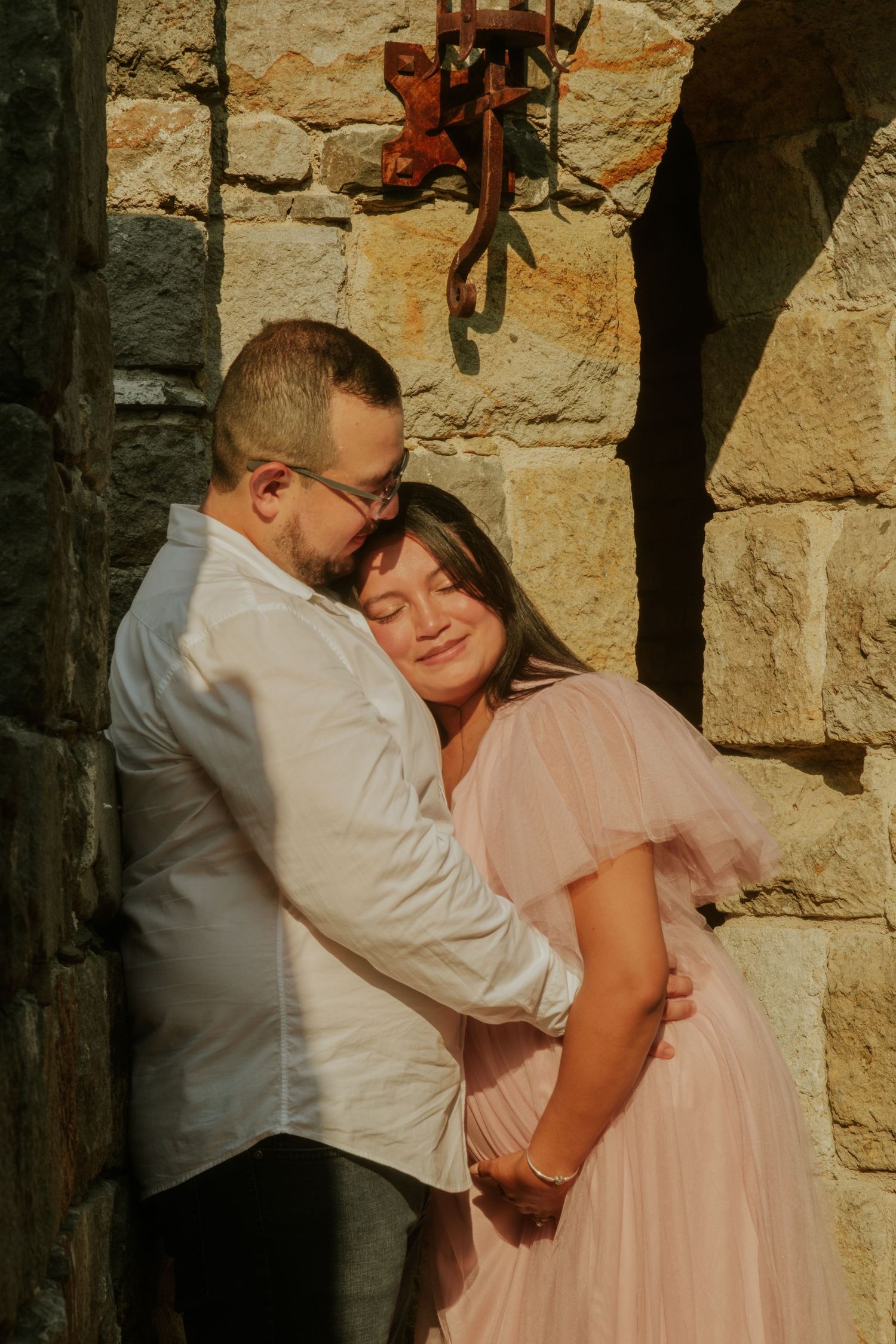 A romantic couple embraces against a rustic stone wall in soft lighting.