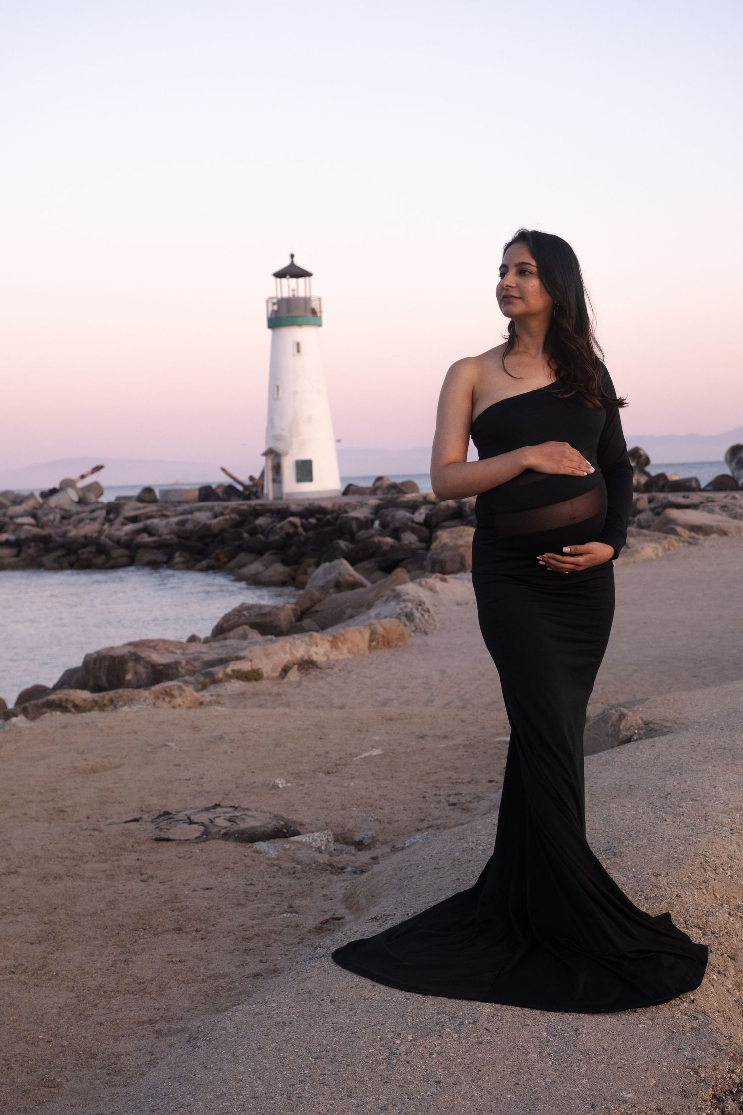 A maternity photoshoot near a white lighthouse on a rocky beach at dusk.