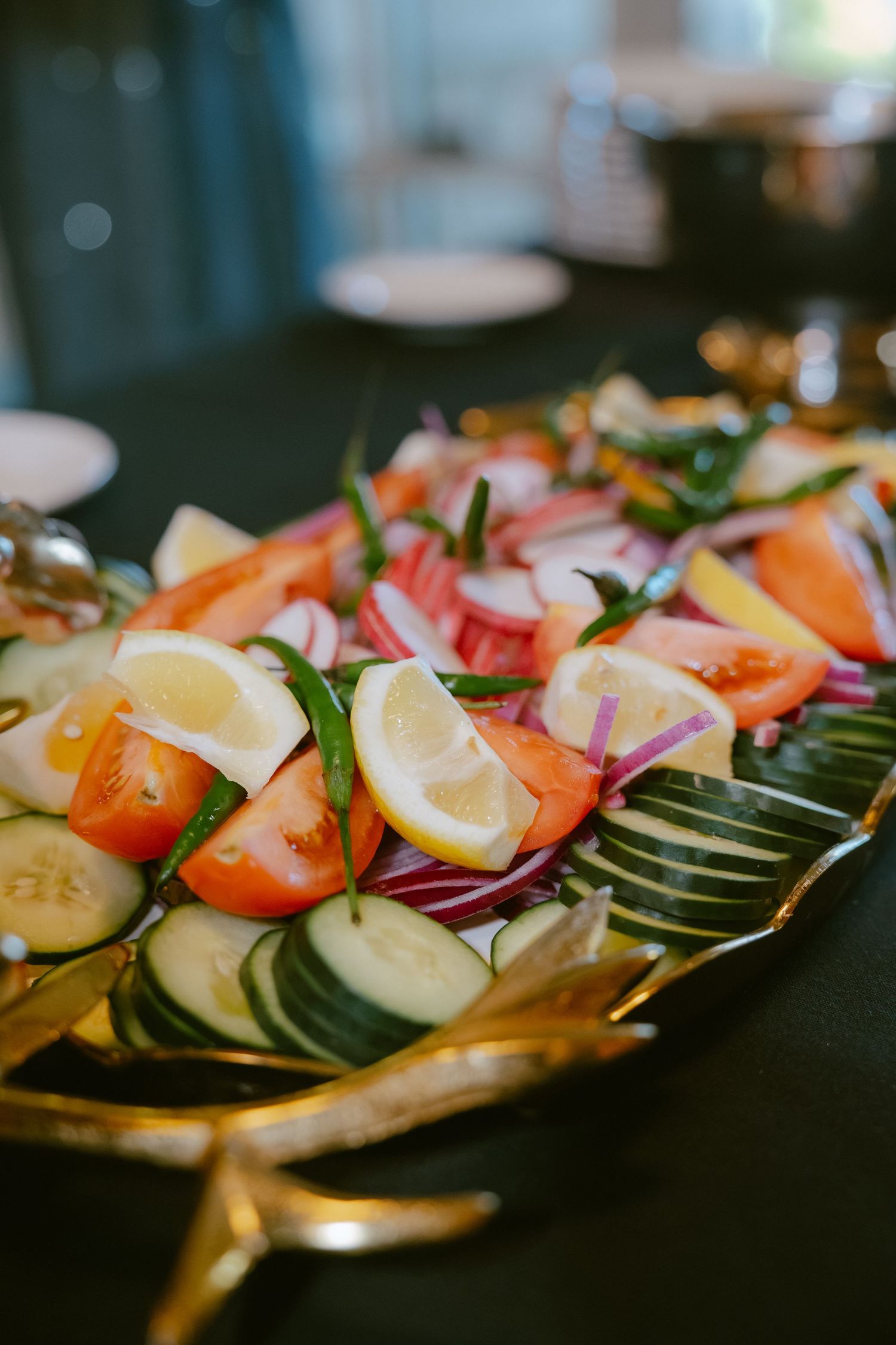Fresh vegetable platter with sliced cucumbers, tomatoes, lemon wedges, and red onions on a gold serving tray.