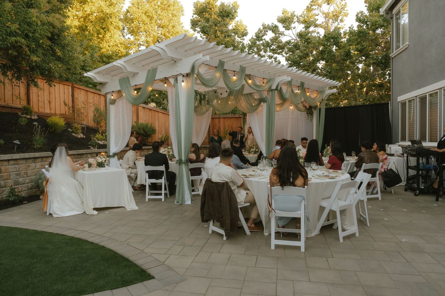 Outdoor backyard wedding reception with guests dining under a white pergola with greenery.