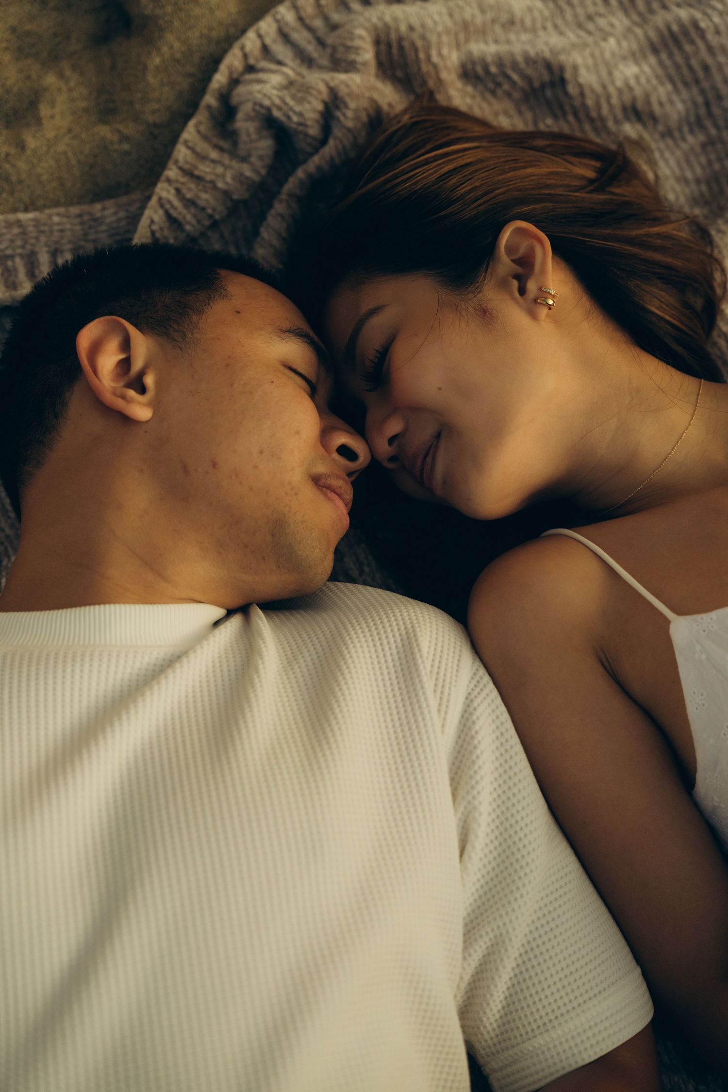 Young couple lying face to face on bed, smiling intimately with eyes closed, wearing white tops.