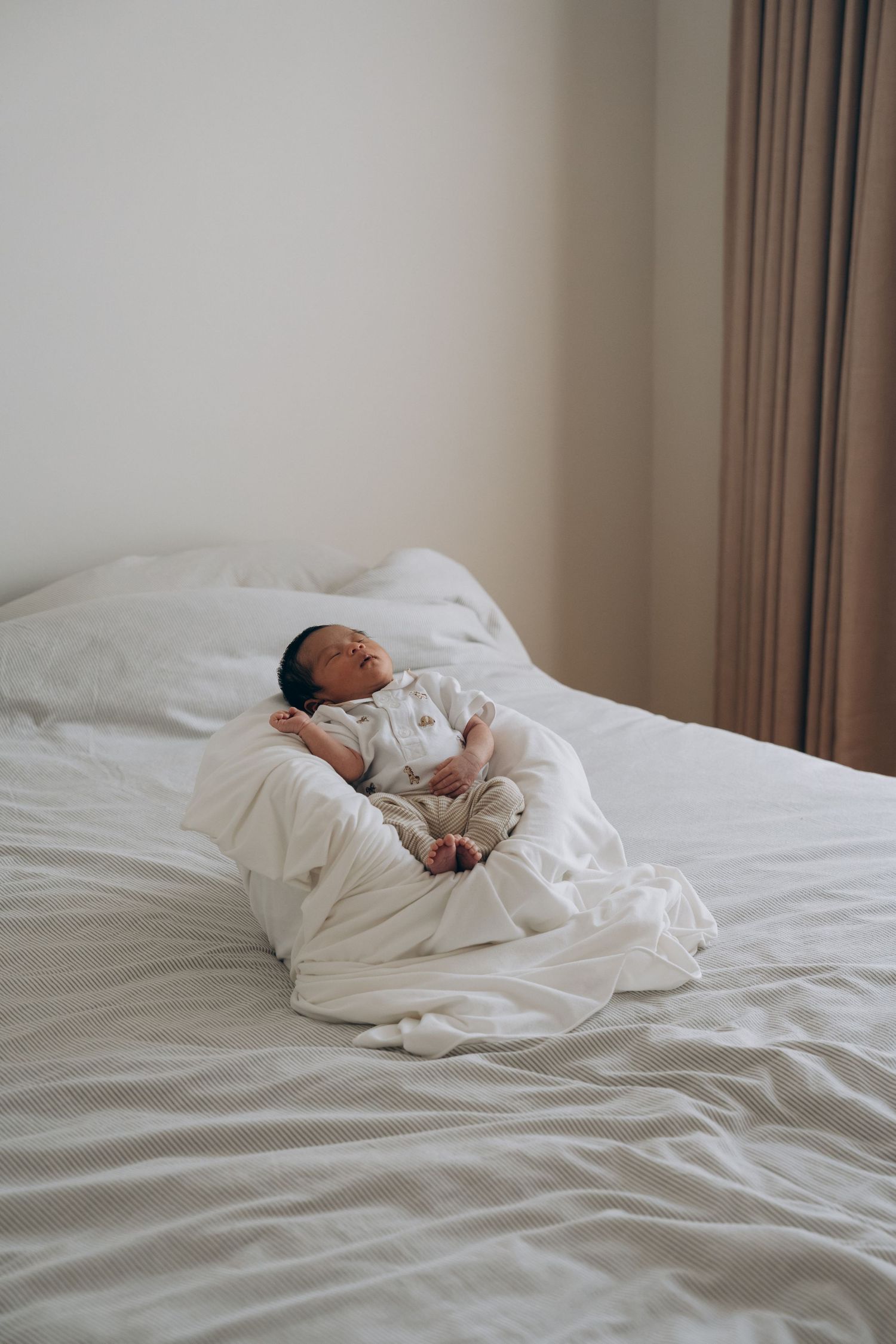 Newborn baby lying awake on a white bed surrounded by soft white blankets in a bright minimalist room.