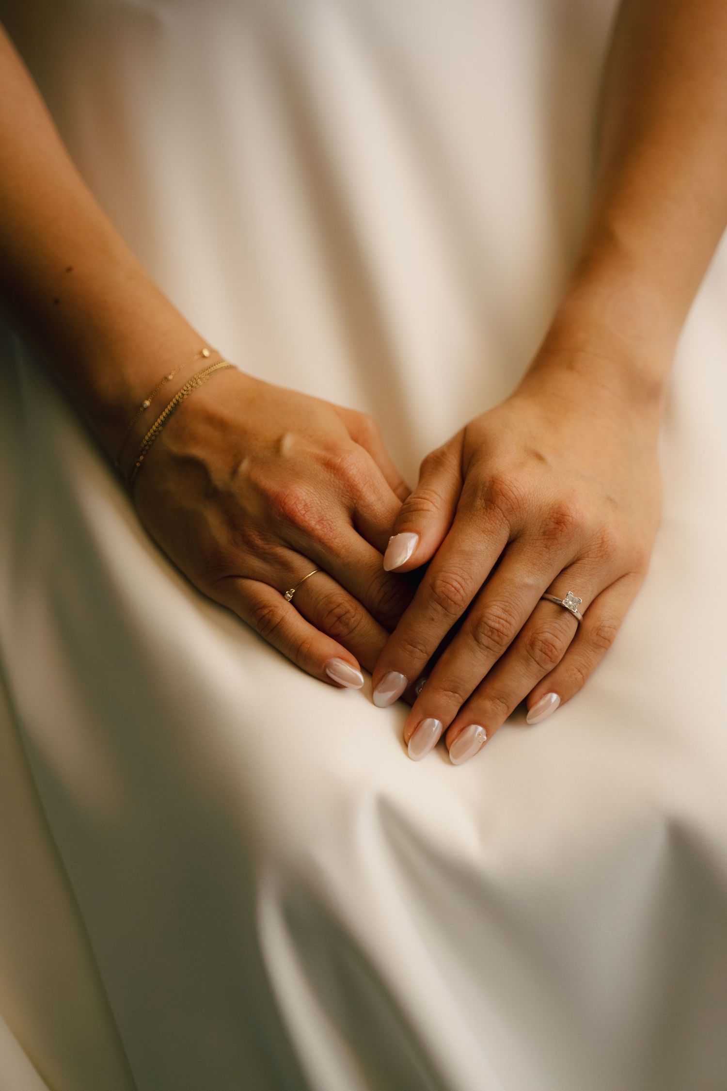 Bride's hands with French manicure, diamond engagement ring, and gold bracelet resting on white wedding dress.
