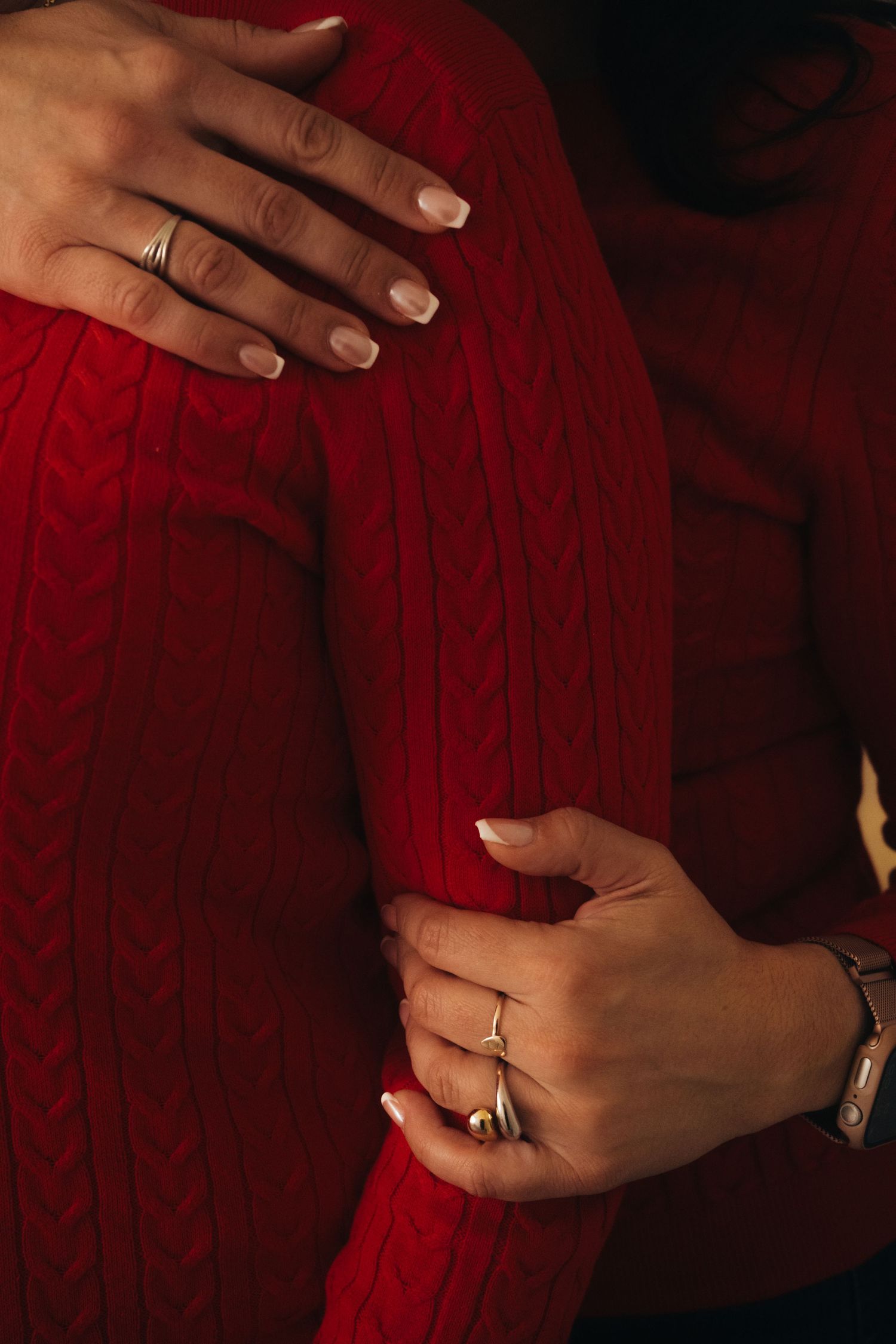 Close-up of hands with rings resting on red cable knit sweater showing intimate holiday moment.