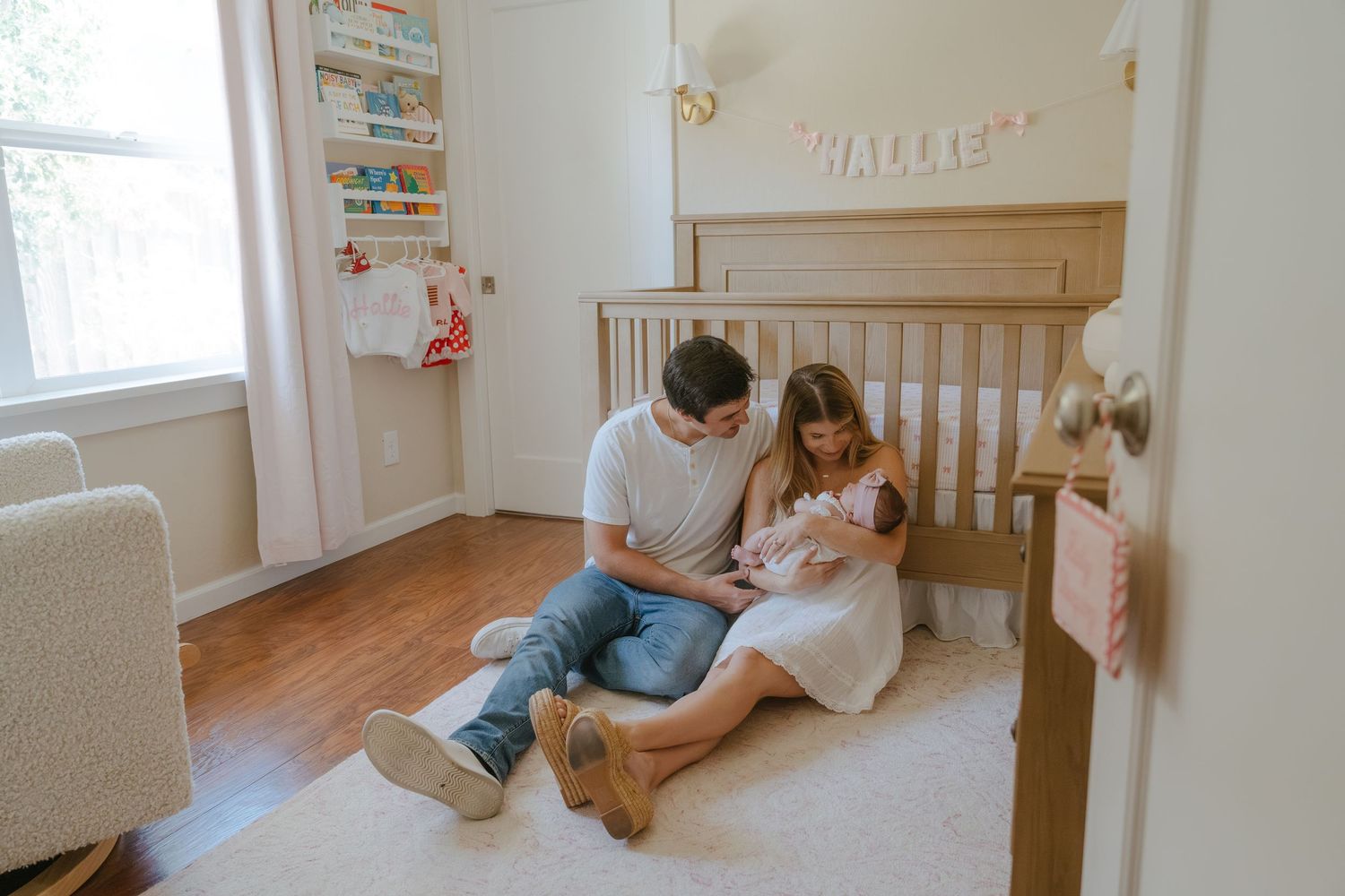 Parents sit on nursery floor with newborn baby near wooden crib, soft rug, and name banner spelling Hallie.