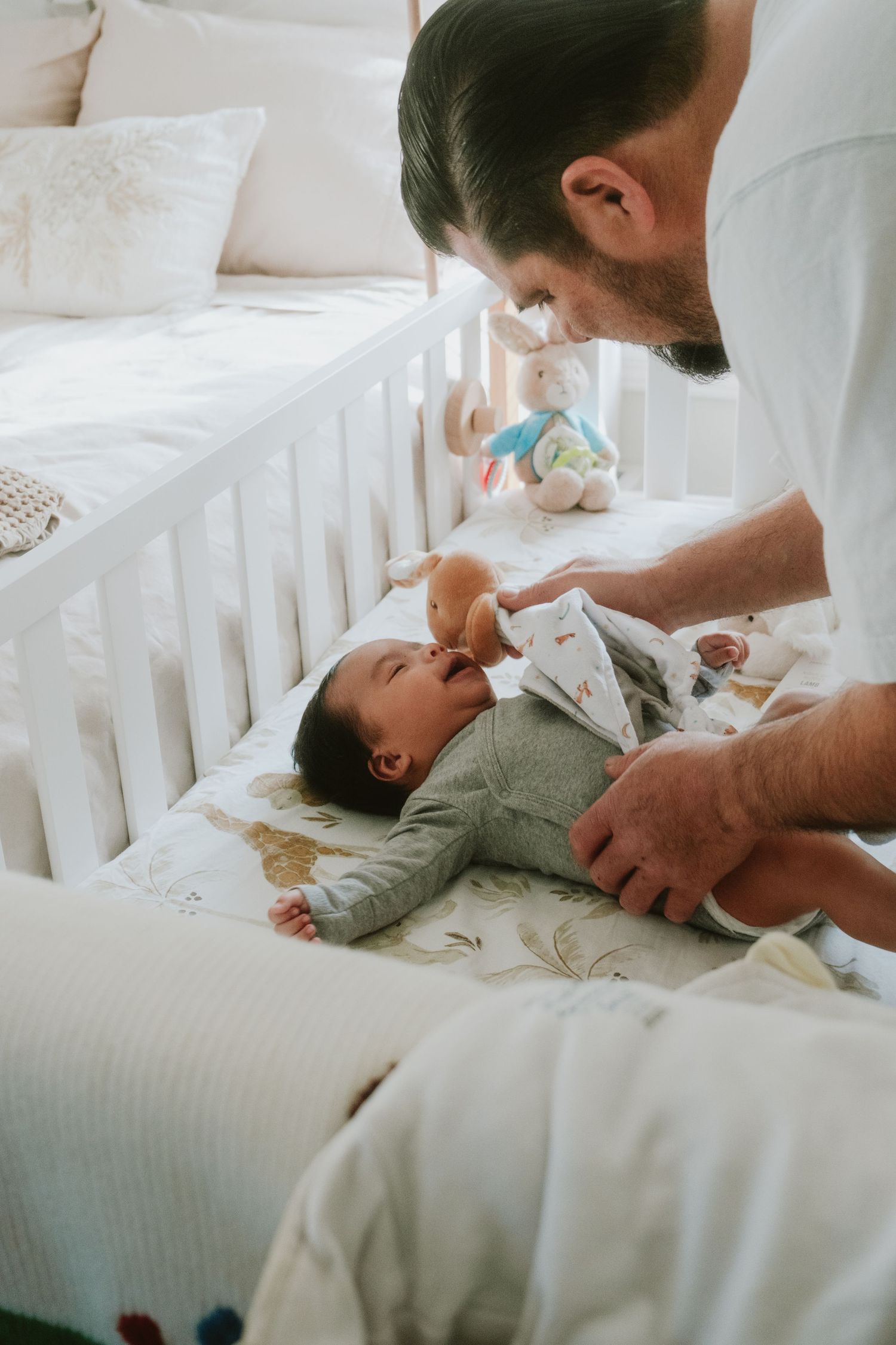 Father smiling at laughing newborn baby in white crib with stuffed animals, tender bonding moment at home.