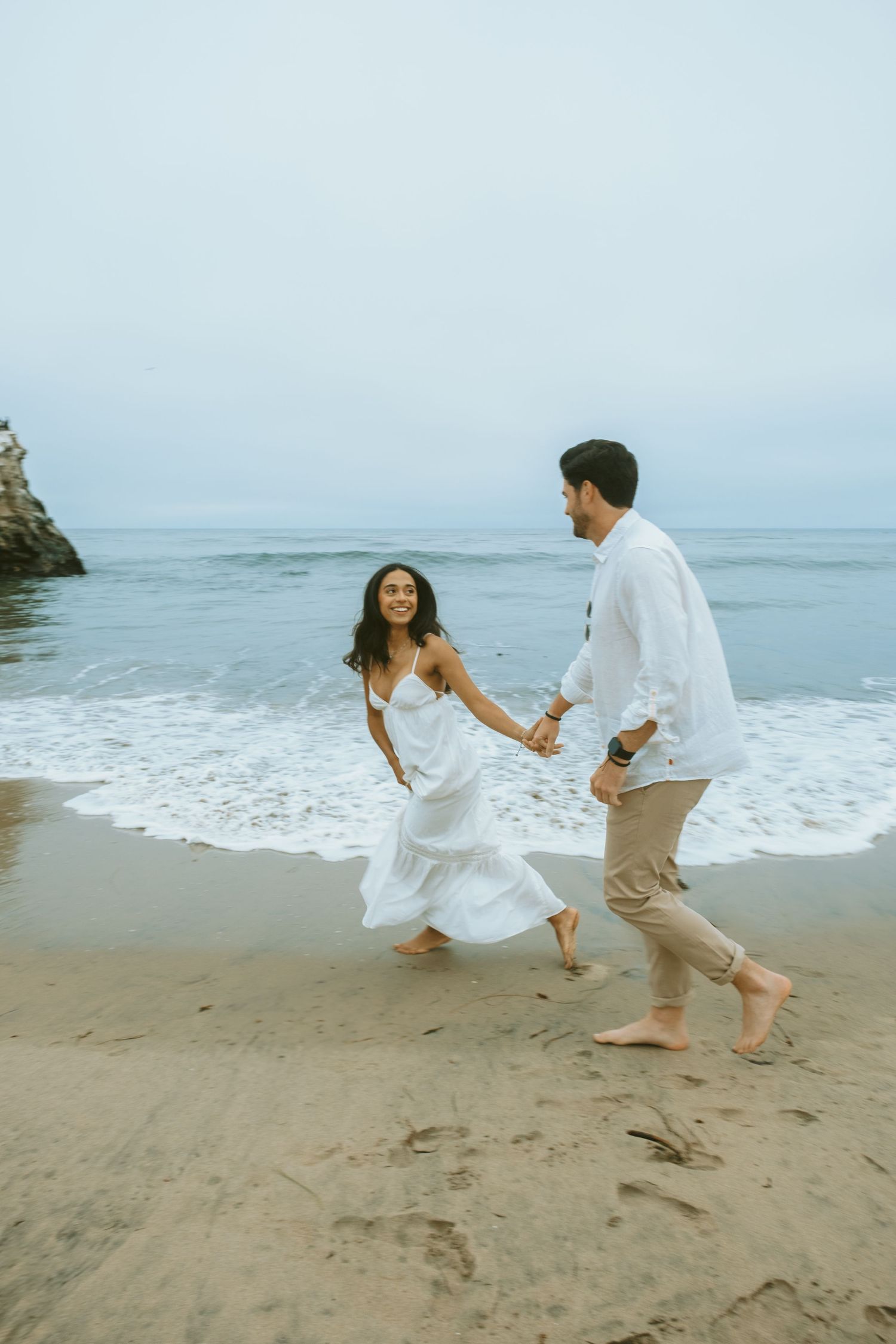 A couple in white outfits holds hands while running barefoot along a sandy beach with ocean waves behind them.
