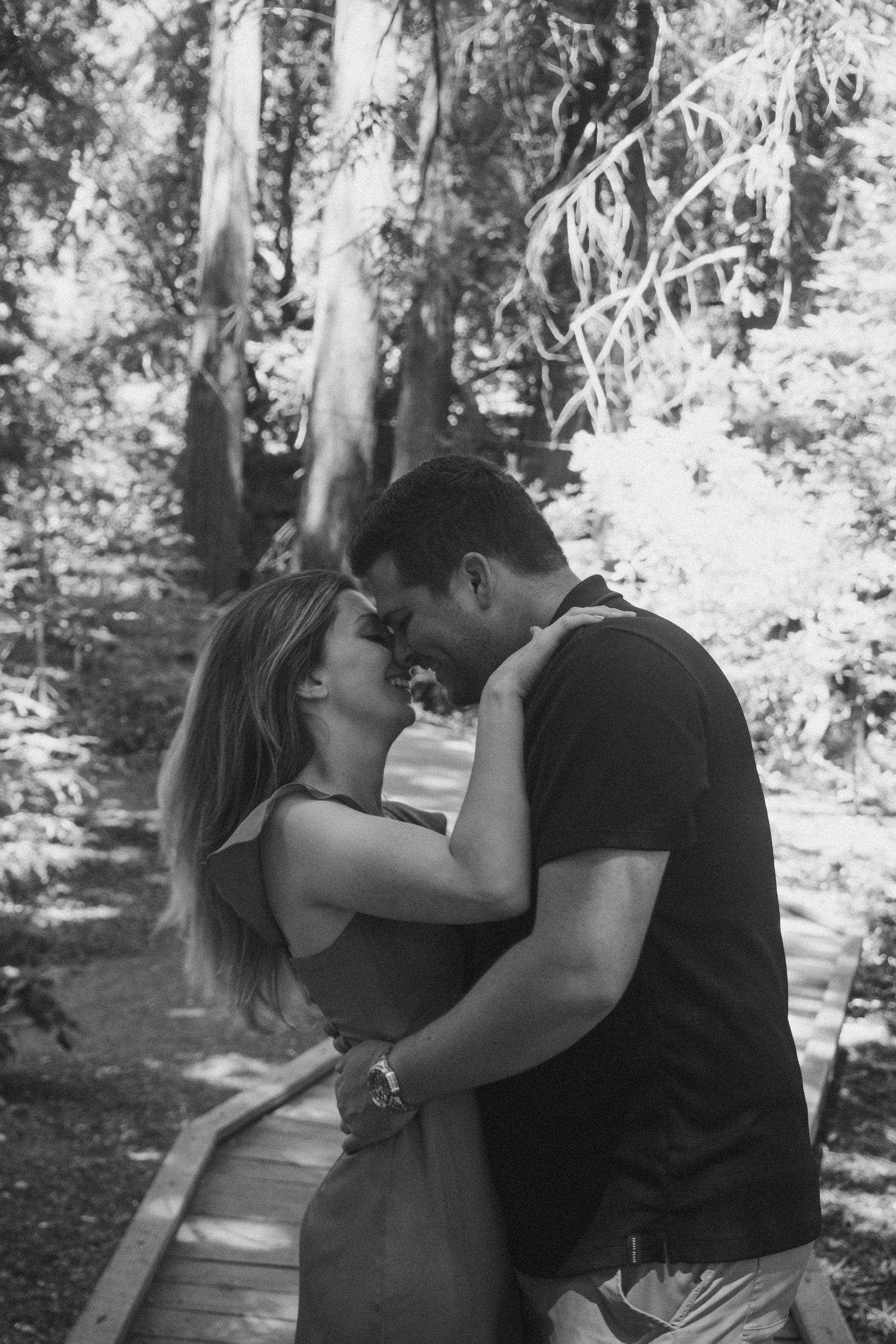 Black and white photo of a couple embracing and about to kiss on a wooden path surrounded by trees.