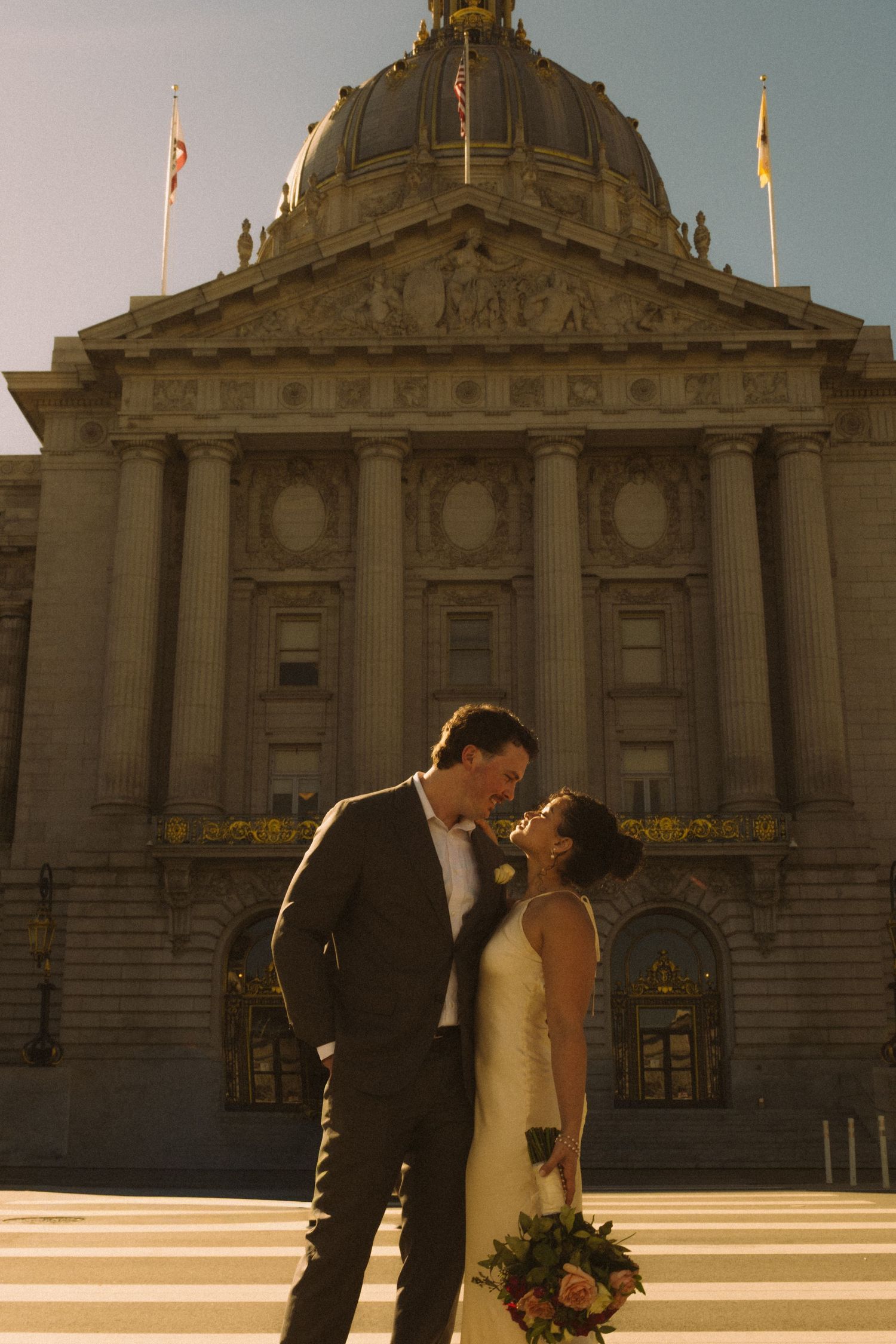 Bride and groom kiss outside San Francisco City Hall at golden hour, bride holding a colorful bouquet.