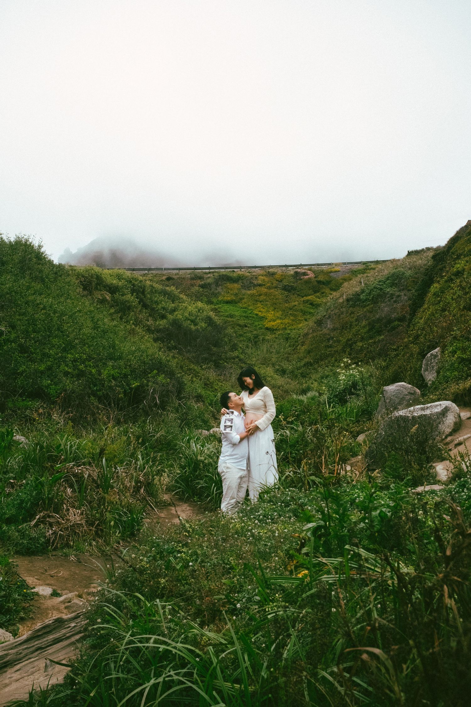 A couple in white attire walks through lush coastal vegetation on a misty day.