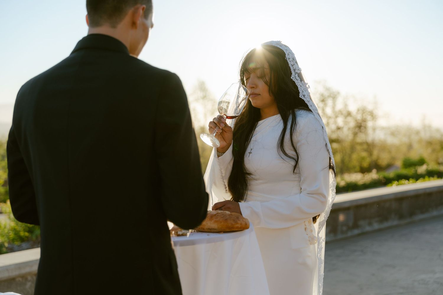 Bride in white dress and veil holding glasses with groom in black suit during outdoor sunset wedding ceremony.