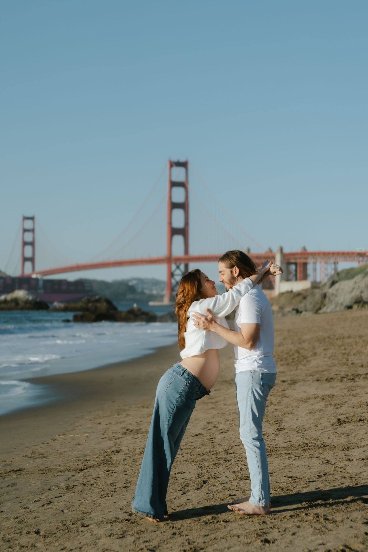 Couple embracing on Baker Beach with the iconic Golden Gate Bridge in the background on a sunny day.