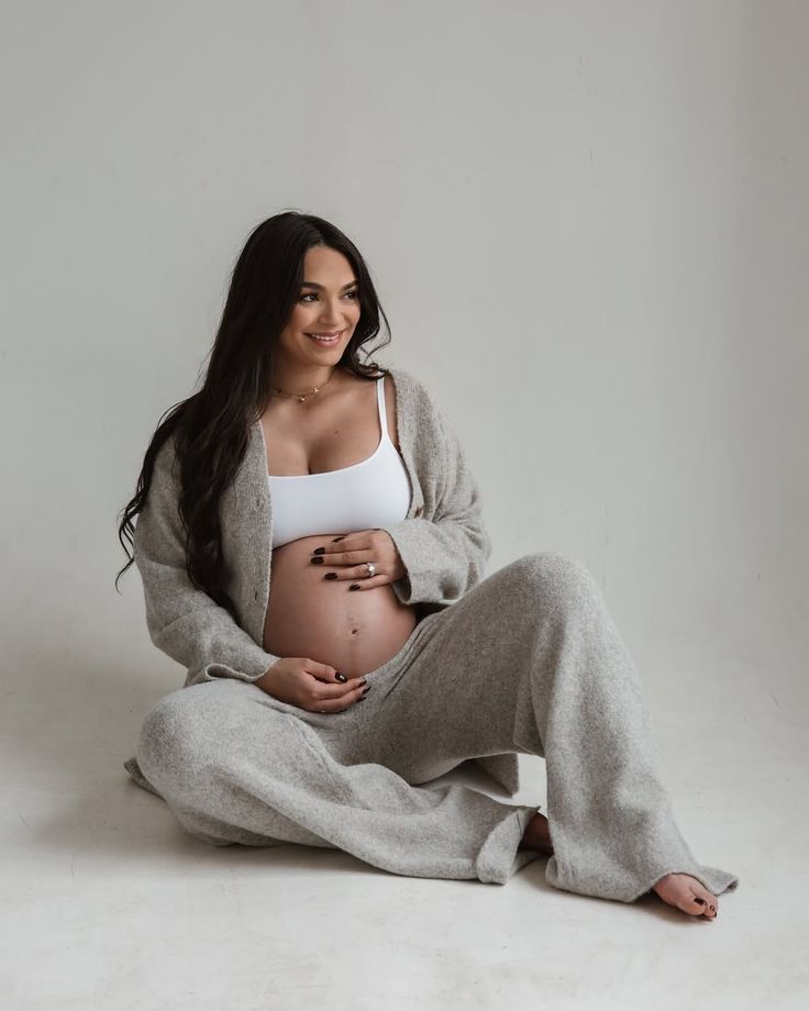 Minimalist maternity photos with woman in cozy gray sweater against white background.