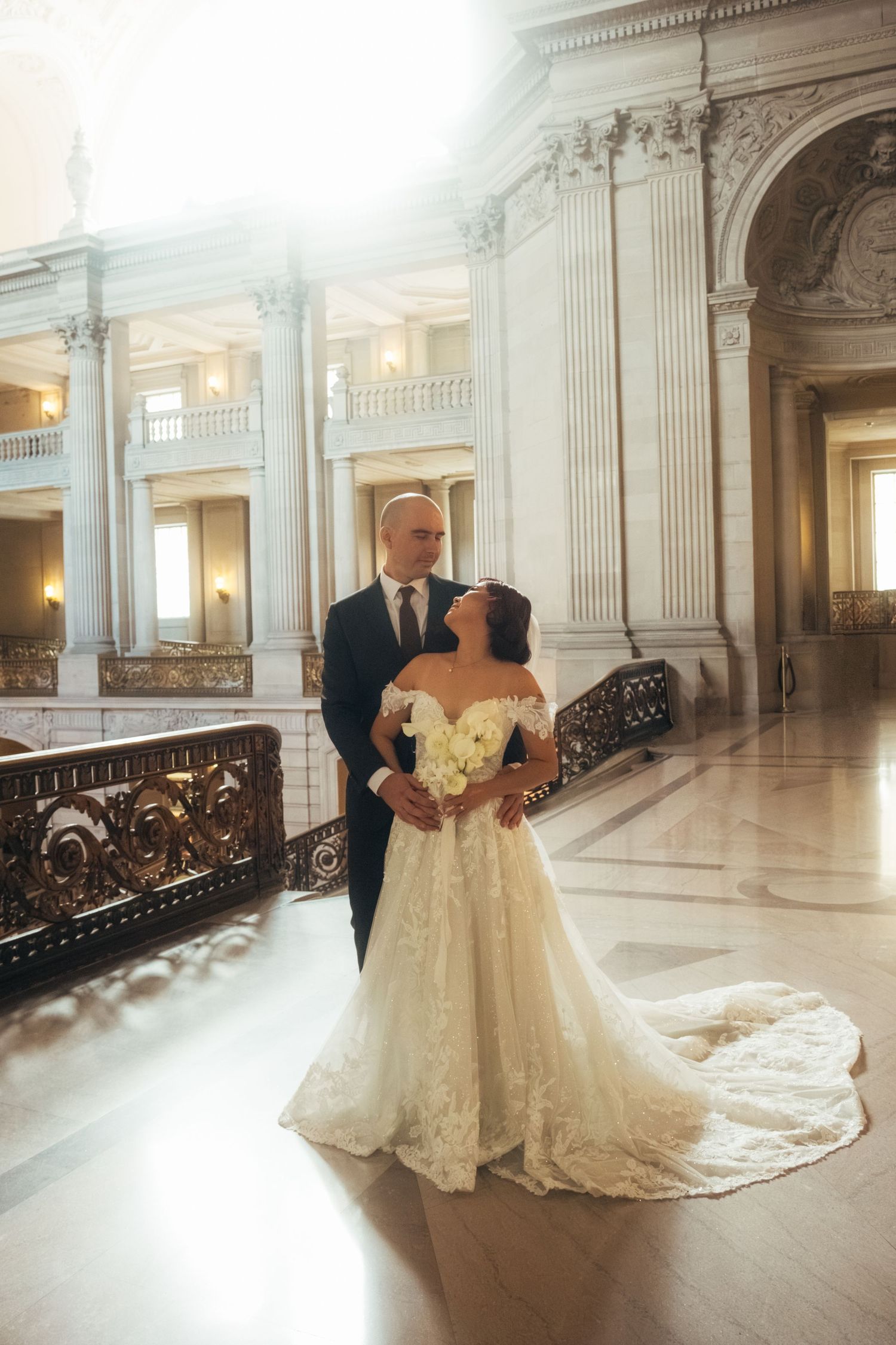 Bride in flowing lace gown and groom embrace in a sunlit classical marble hall.