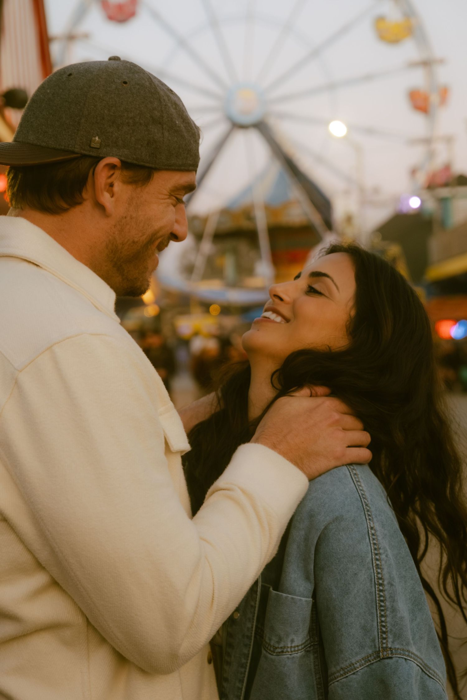 Happy couple sharing a romantic moment at a colorful carnival with a Ferris wheel in the background.