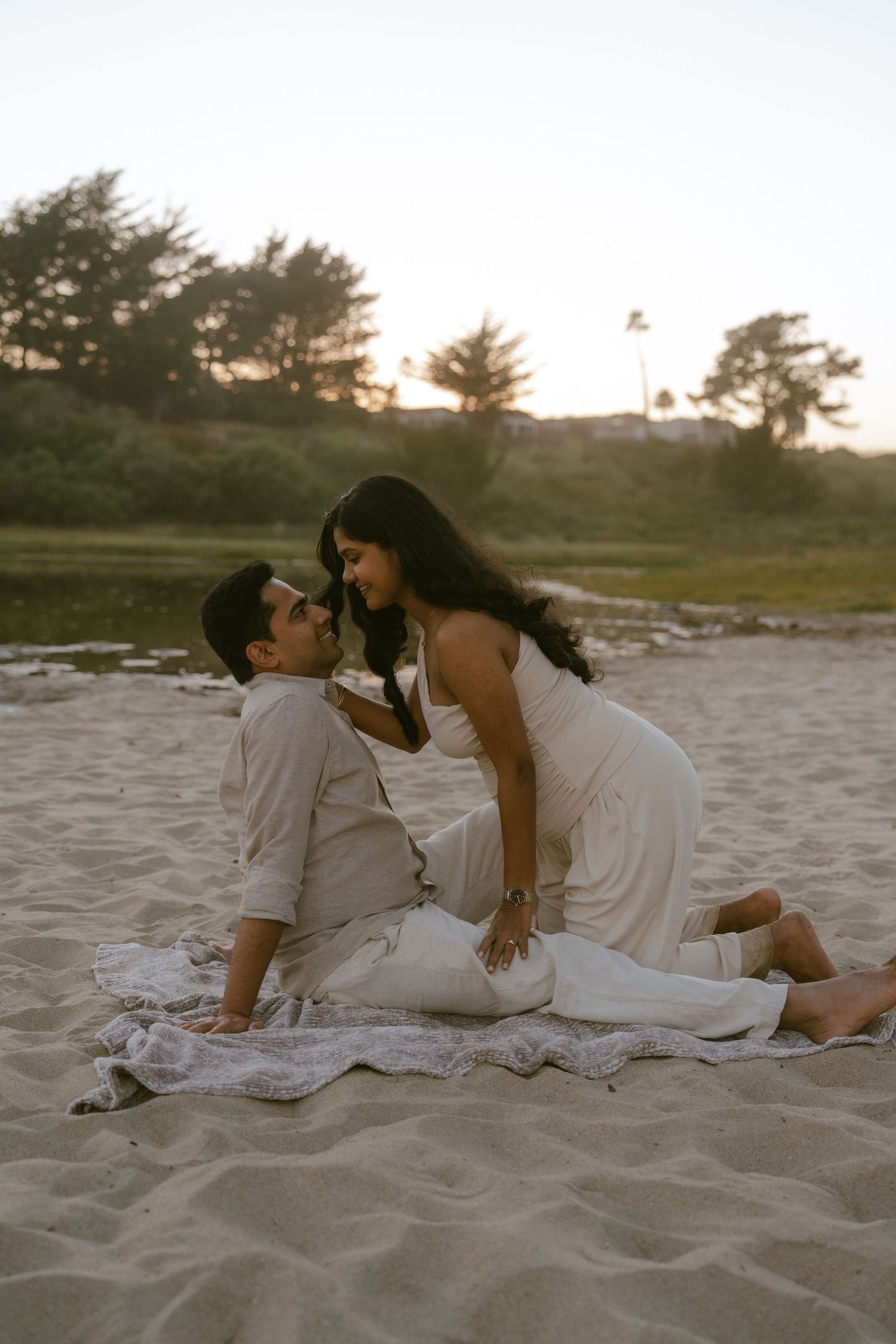 Couple in white outfits sharing a romantic moment on sandy beach at sunset, sitting on a blanket.