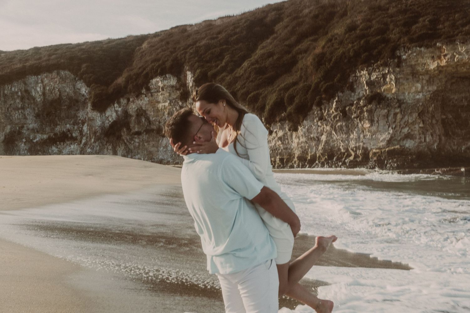 Couple sharing a romantic kiss on a scenic beach with ocean waves and dramatic cliffs in the background.