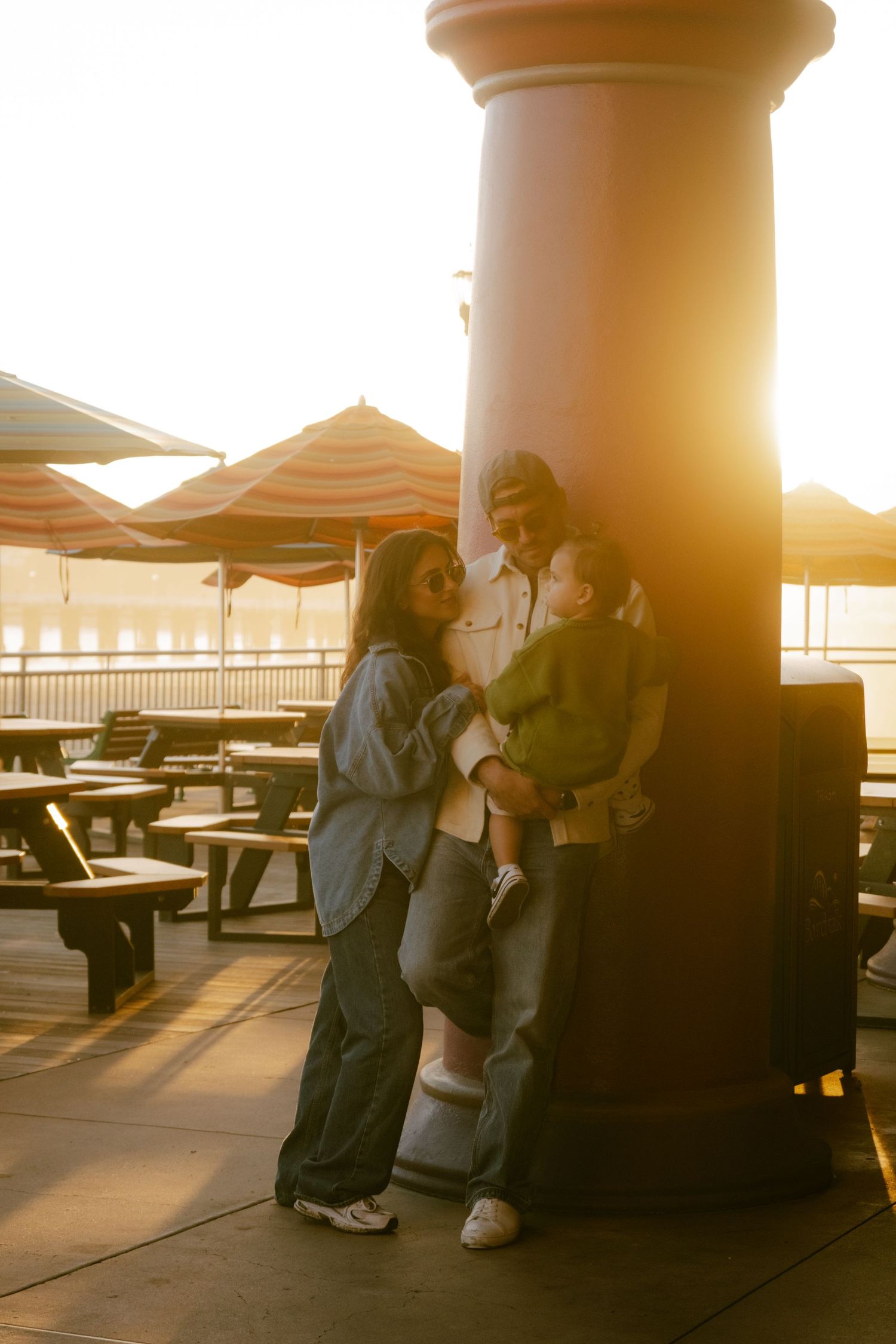 Family of three posing by a pillar at sunset on a waterfront patio with striped umbrellas in the background.