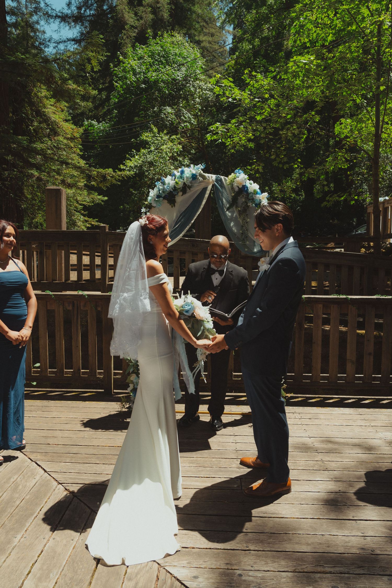 Bride and groom exchange vows on a wooden deck surrounded by lush trees, with an officiant and floral arch behind them.