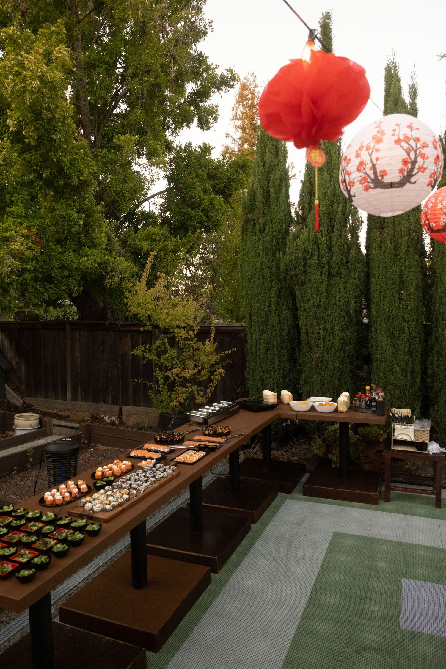 Outdoor sushi buffet table with paper lanterns hanging above, set in a garden with cypress trees.