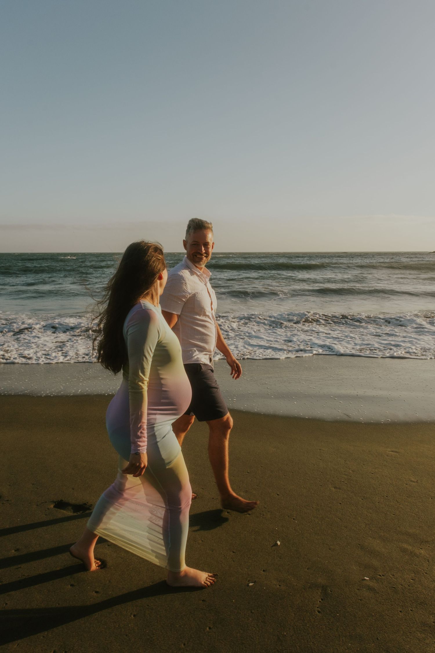 A couple walking along a sandy beach at sunset with waves crashing behind them.