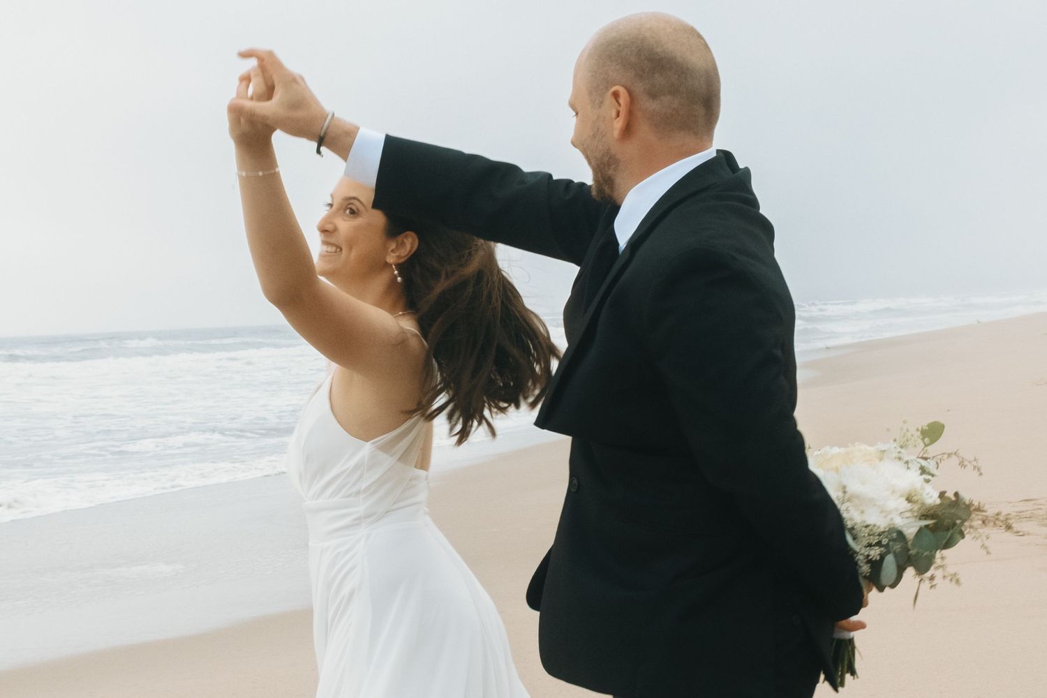 Wedding couple sharing an intimate dance moment on a sandy beach with waves behind them.