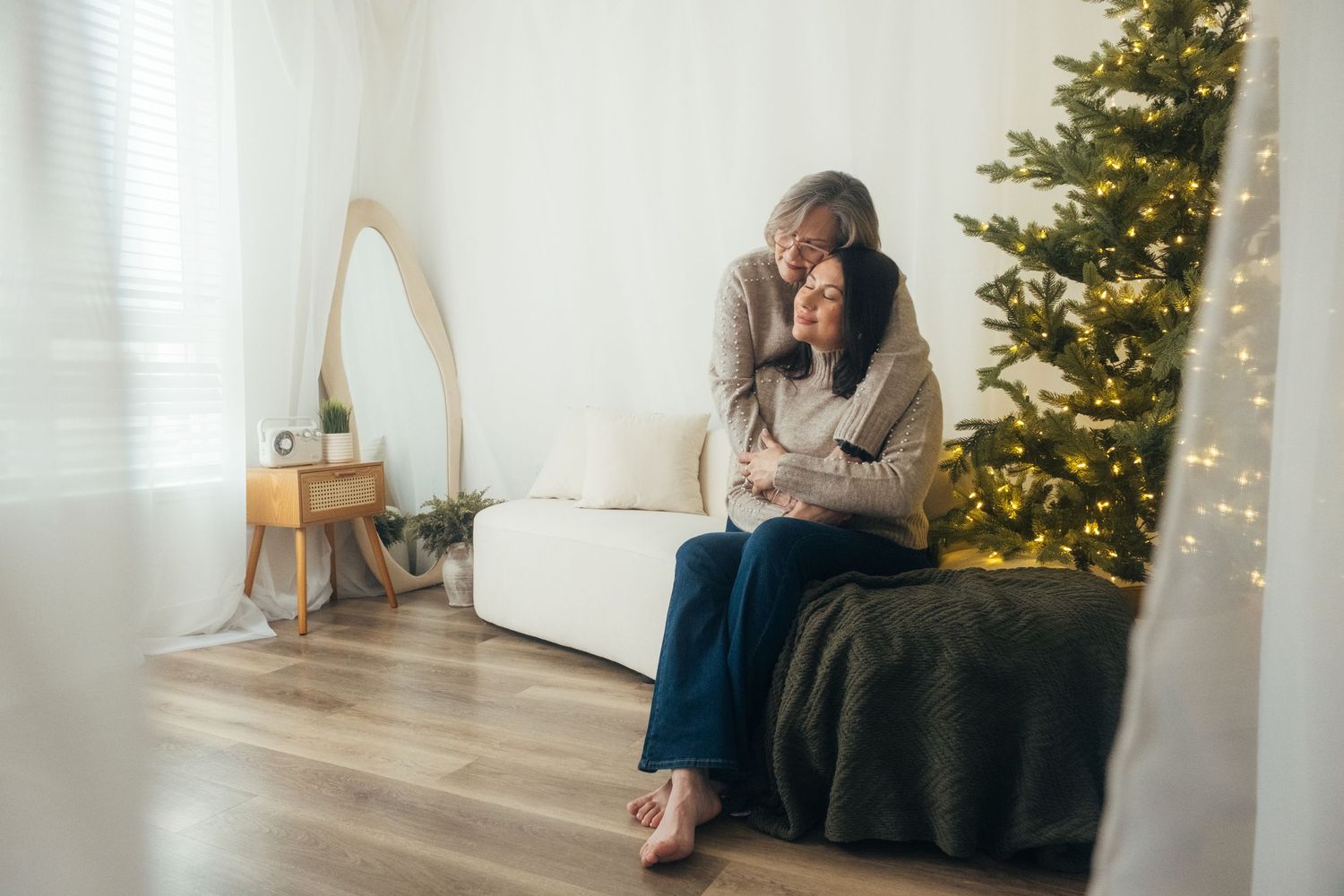 Person sitting alone on couch with emotional expression near decorated Christmas tree.