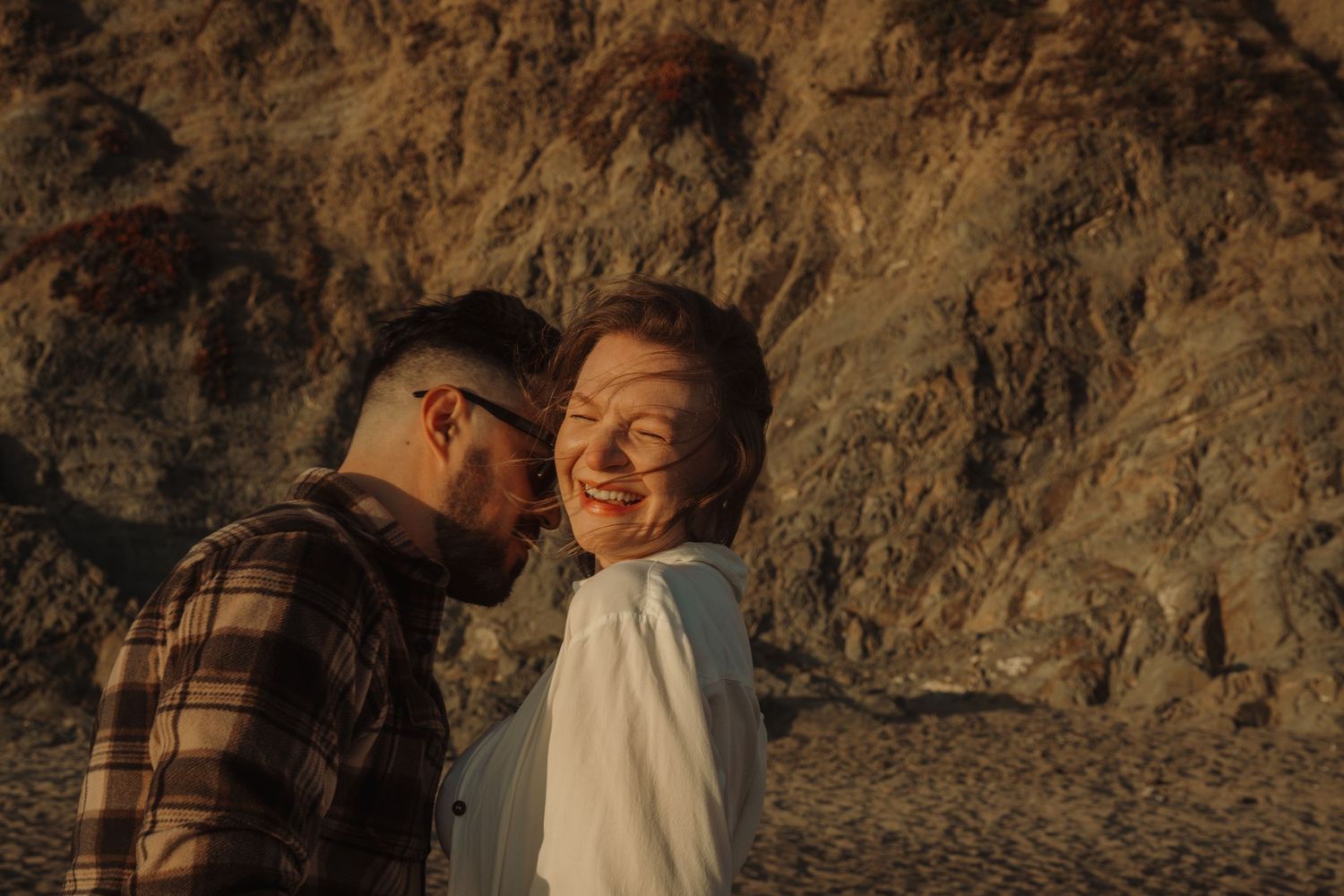 Romantic couple's portrait session against rocky beach cliffs at sunset.